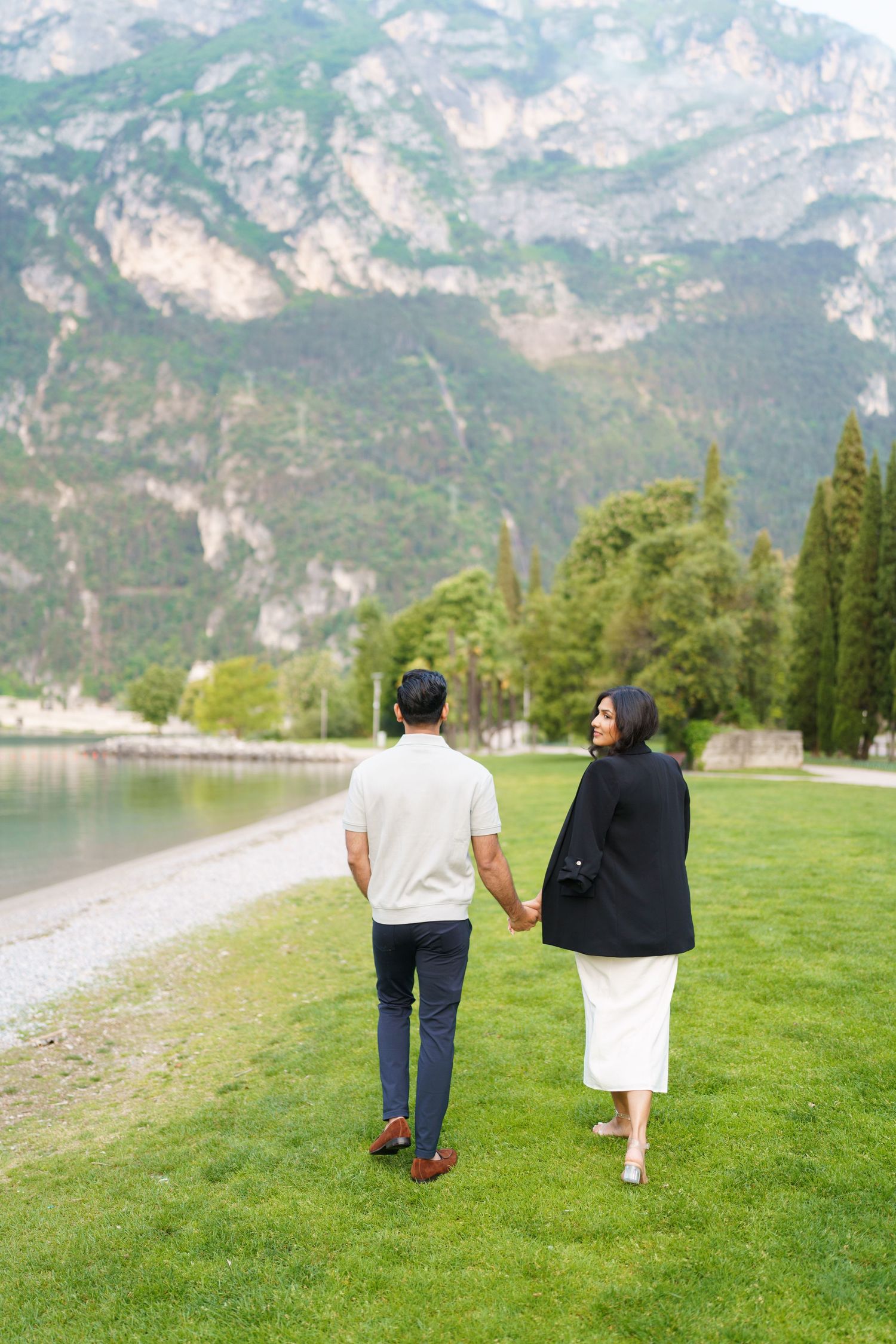 A couple walks hand in hand along a lakeside path with dramatic mountain peaks rising in the background.