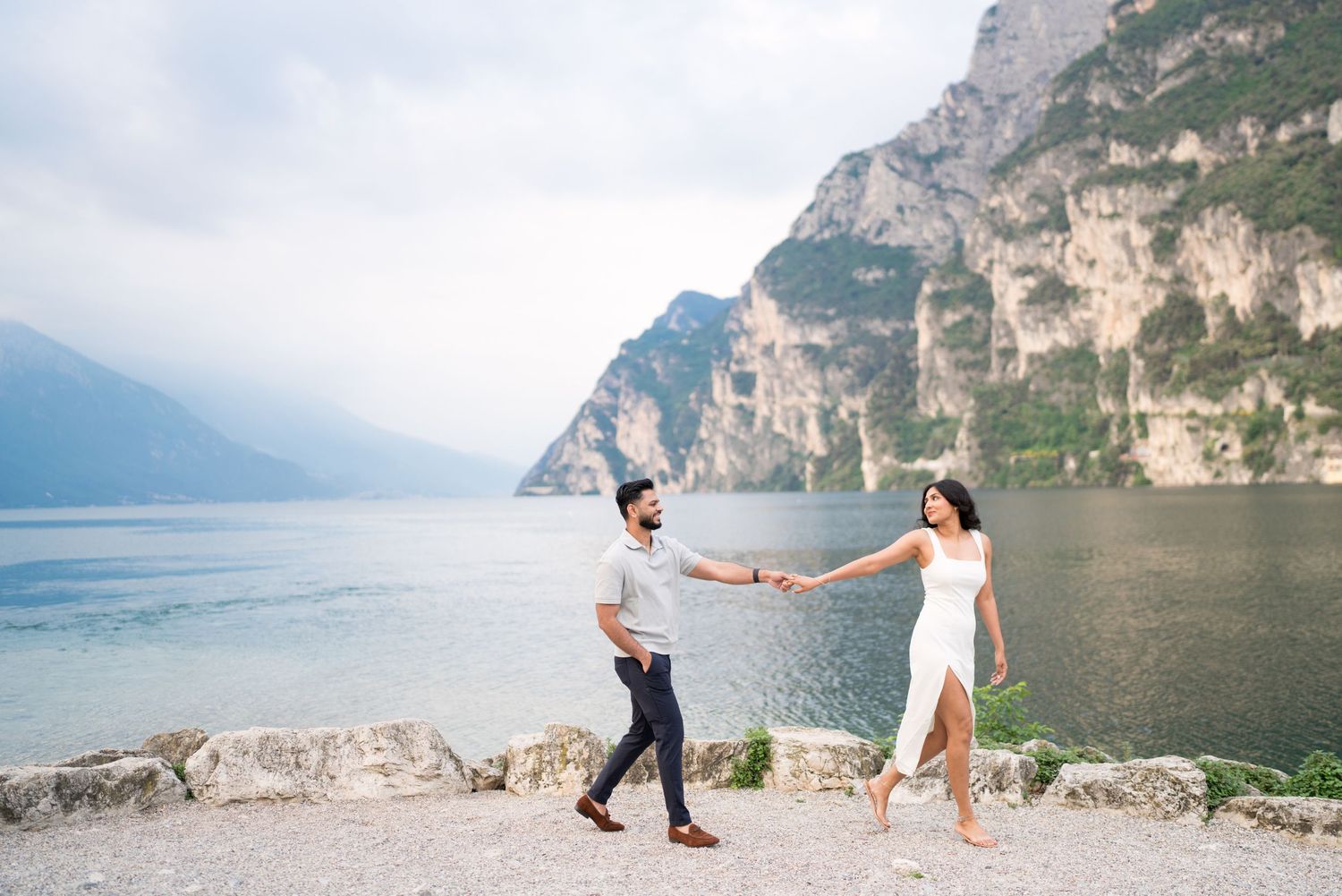 Two people hold hands while dancing together on a scenic mountain path overlooking a lake in Italy.