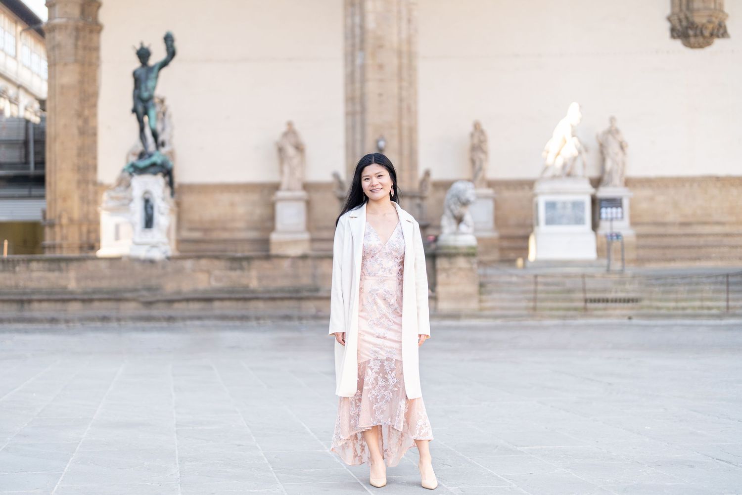 A person in a pink blazer and white dress poses in front of a stone building facade with arches.