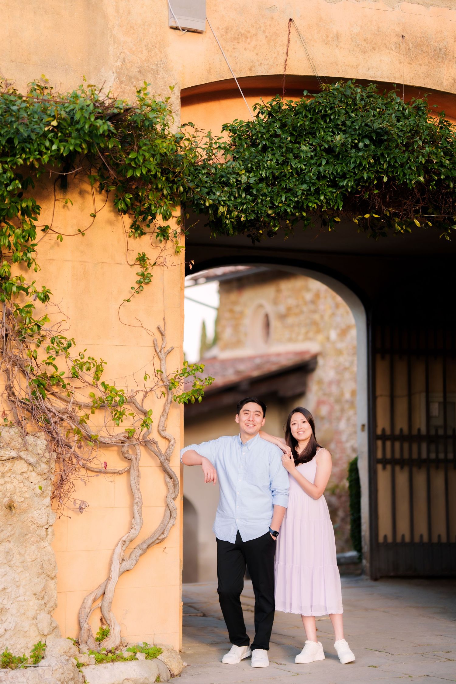 A couple stands under an arched passageway with climbing vines on a peach-colored Mediterranean style wall.