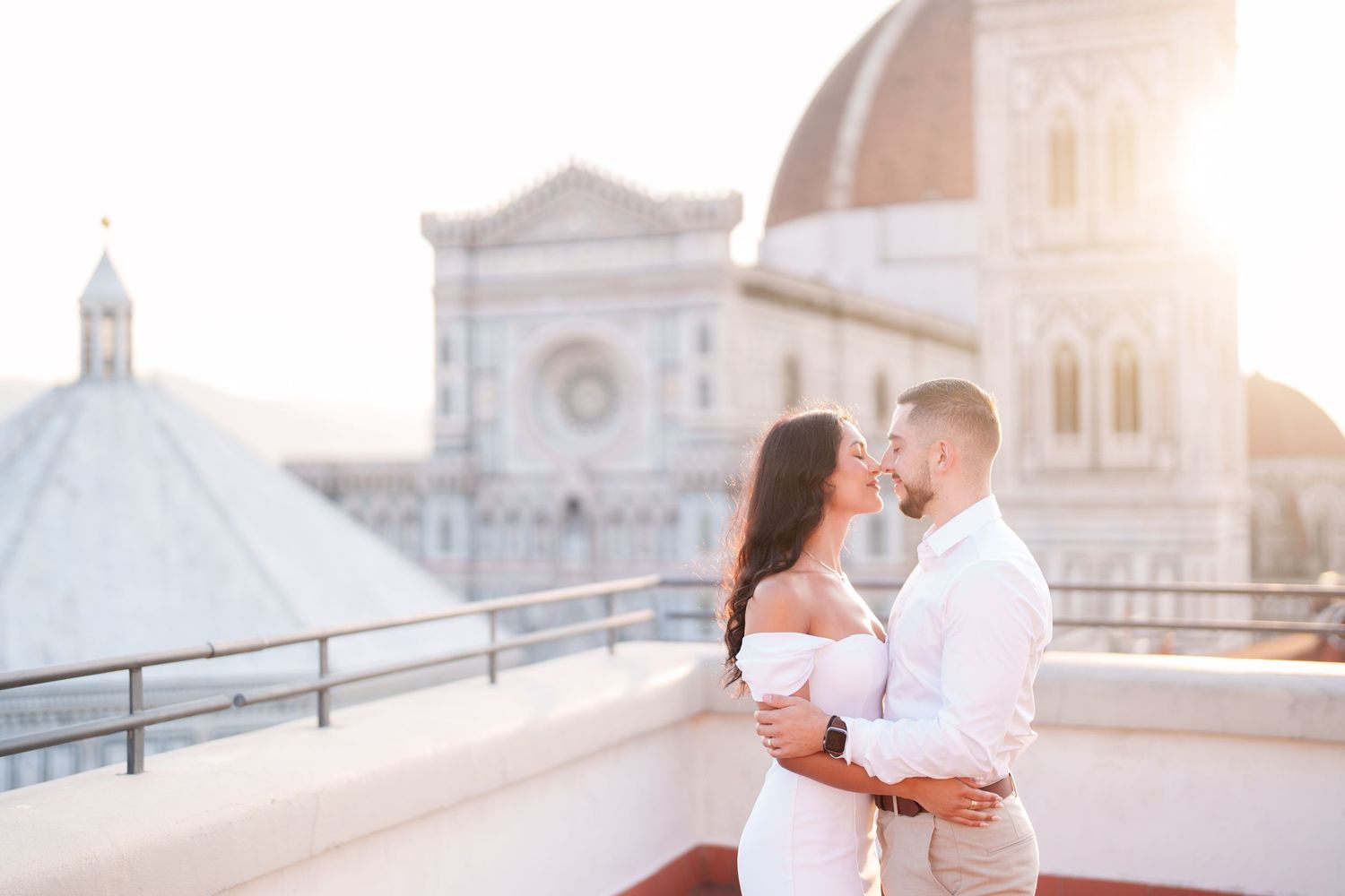 Romantic couple shares intimate moments at sunset on a European city bridge with ornate architecture in the background.