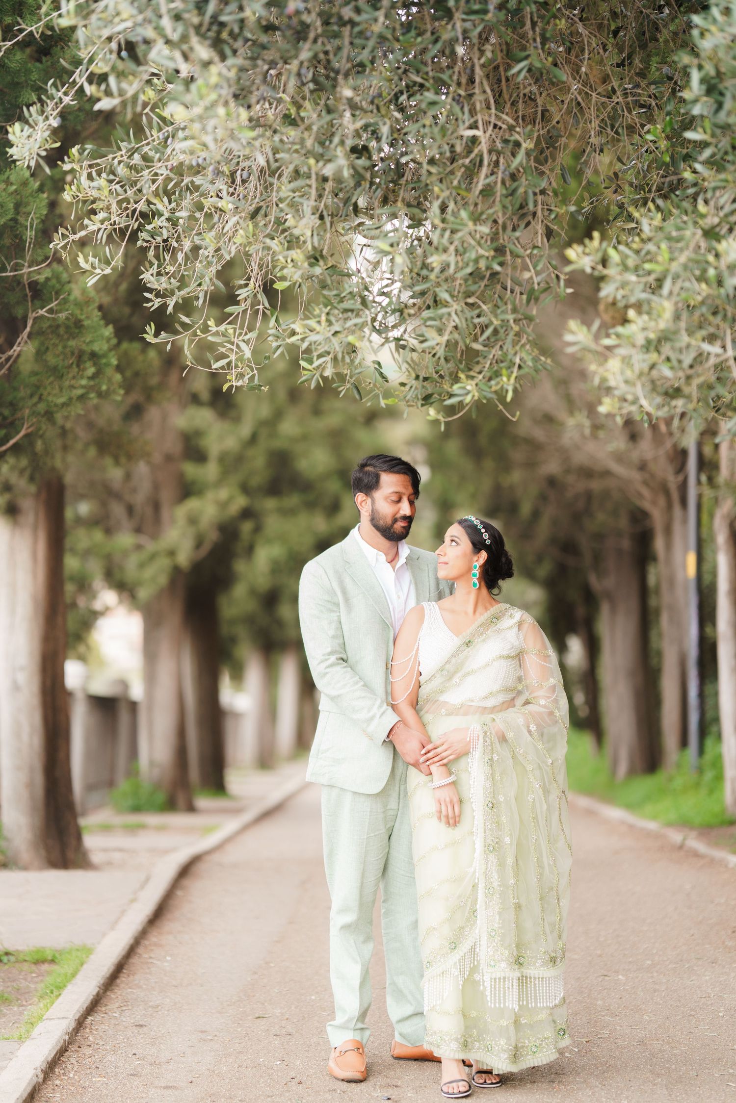 A couple in elegant white and mint green outfits walk hand in hand down a tree-lined path during a romantic photoshoot.