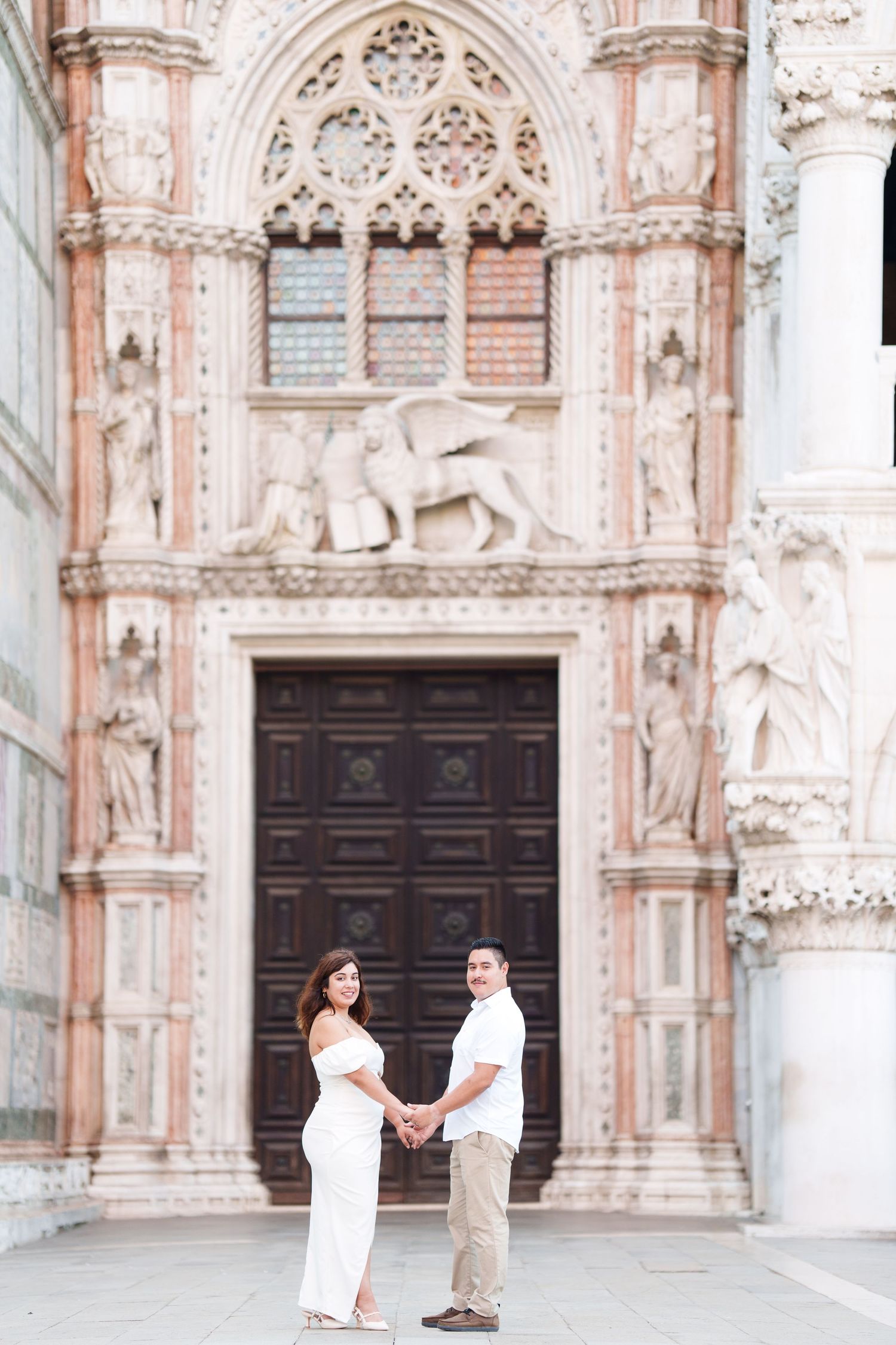 A couple poses for romantic photos in front of an ornate Gothic cathedral with detailed stone carvings and dark wooden doors.