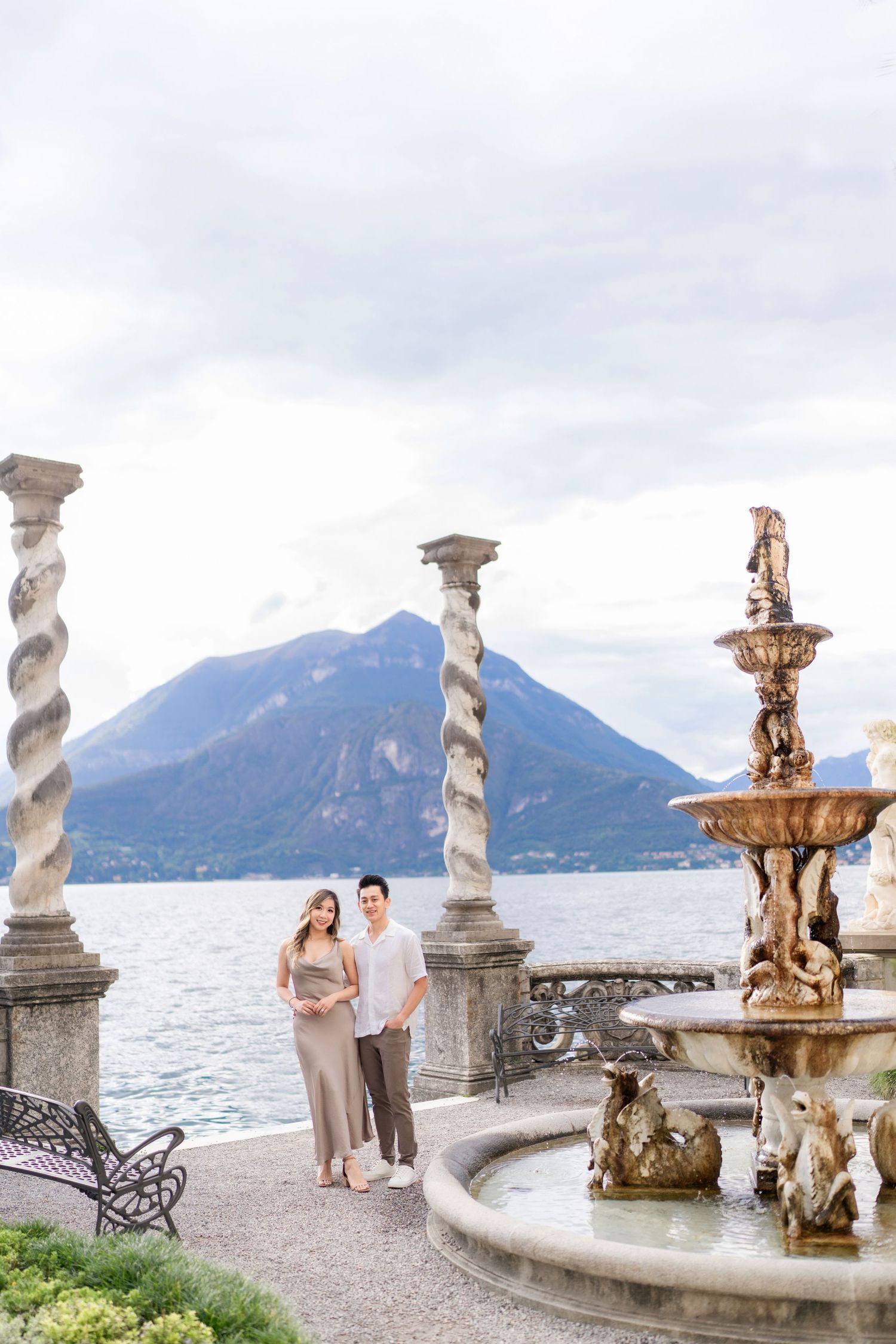 Romantic couple standing by ornate fountain with Lake Como and mountains in background at Villa Monastero, Italy.