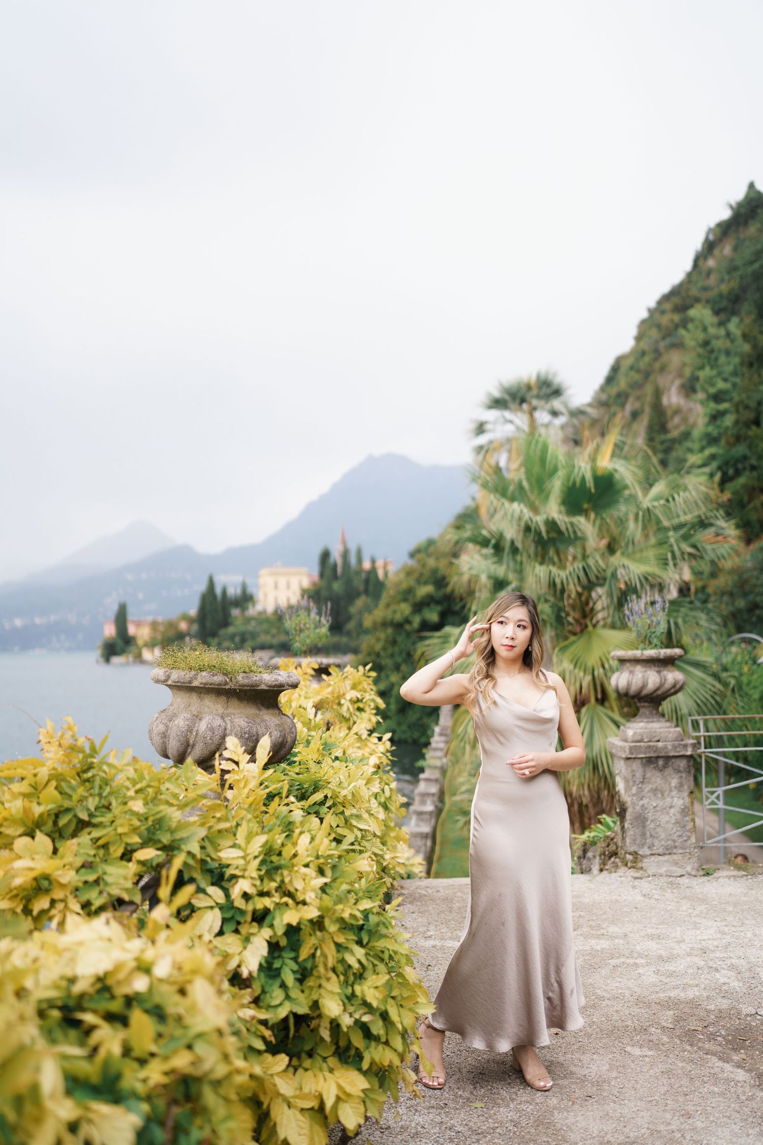 A person in a flowing dress poses in a scenic Mediterranean garden setting with mountains and coastline in the background.