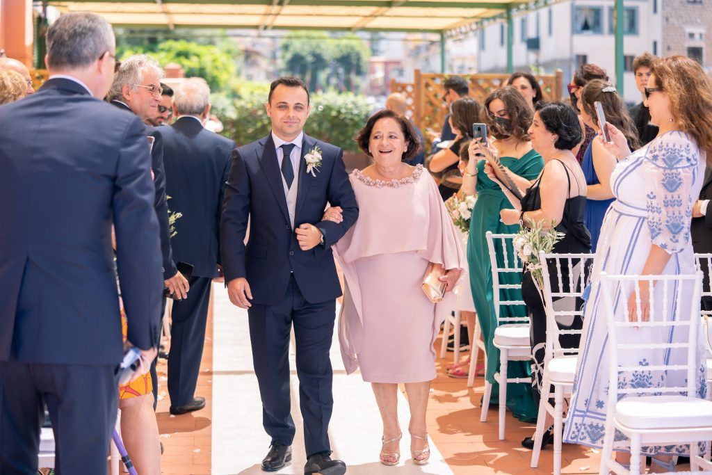 Wedding guests in formal attire gather for outdoor ceremony with white chairs.