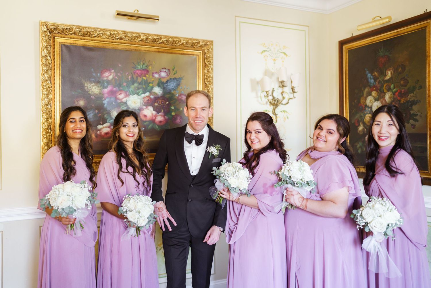 Wedding party in purple bridesmaid dresses and black tuxedos pose in elegant white room.