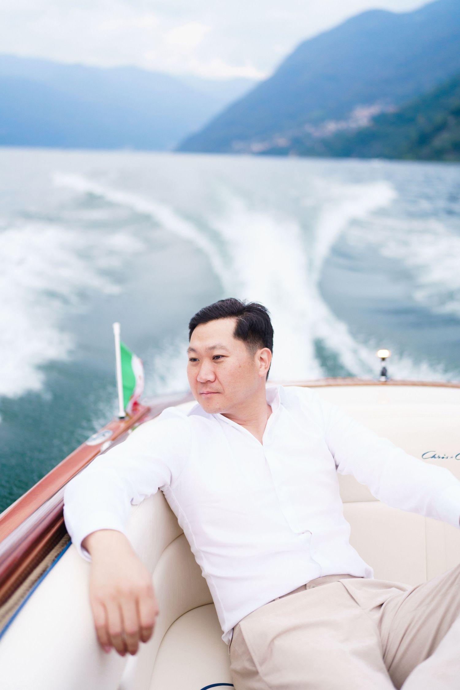 Person in white clothing relaxes on a luxury boat with mountainous coastline in the background.