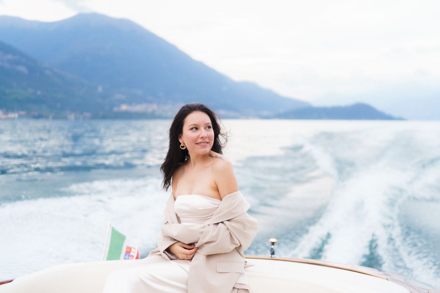 Someone relaxes on a boat wearing a beige sweater with mountains and wake trails visible in the background on a misty day.