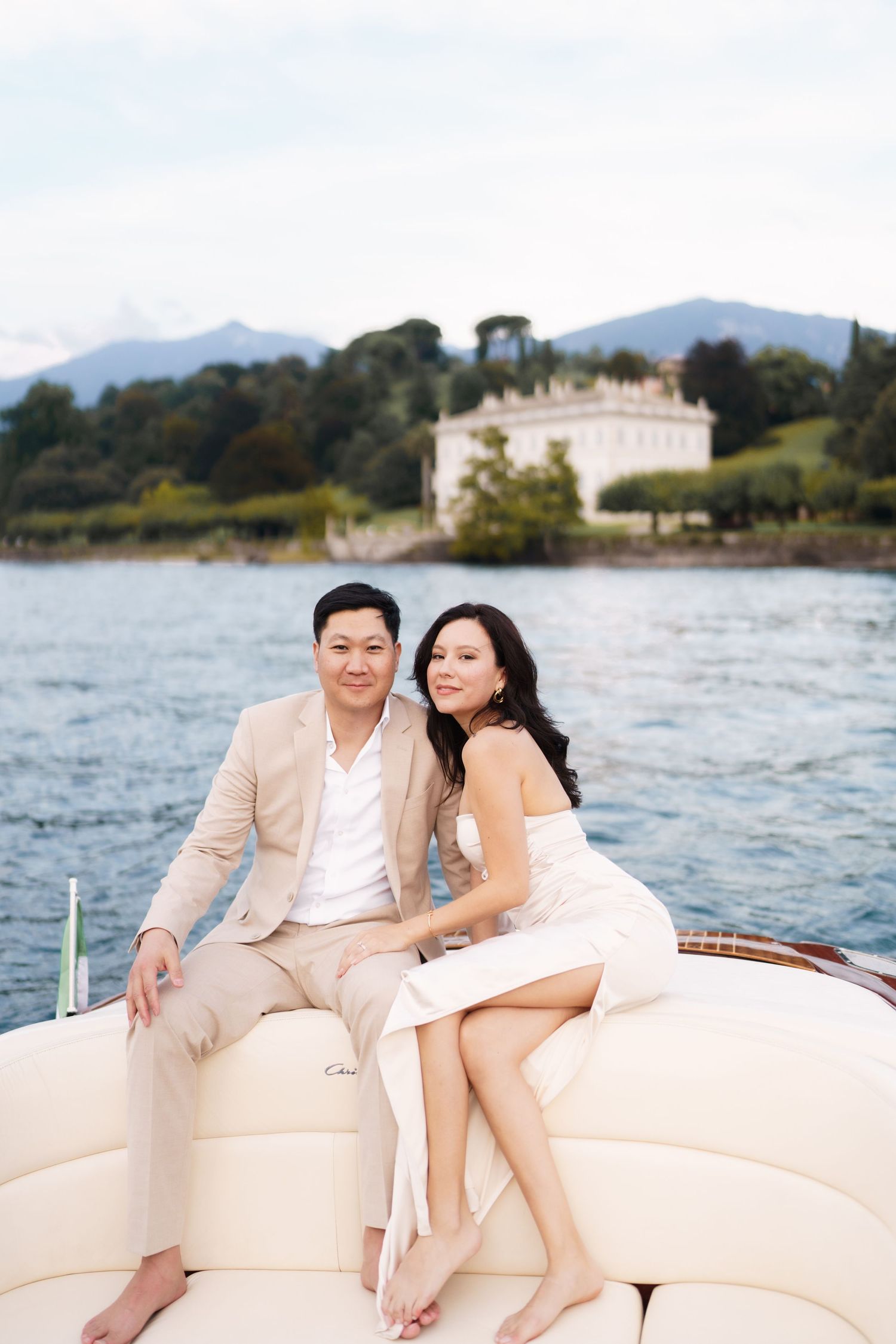 Couple enjoying a romantic boat ride along the scenic lakefront with mountains and villas in the background.
