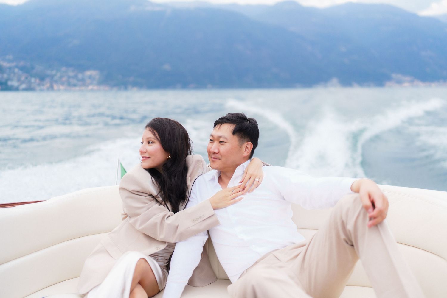 A couple relaxes together on a white boat seat with mountains and water in the background.