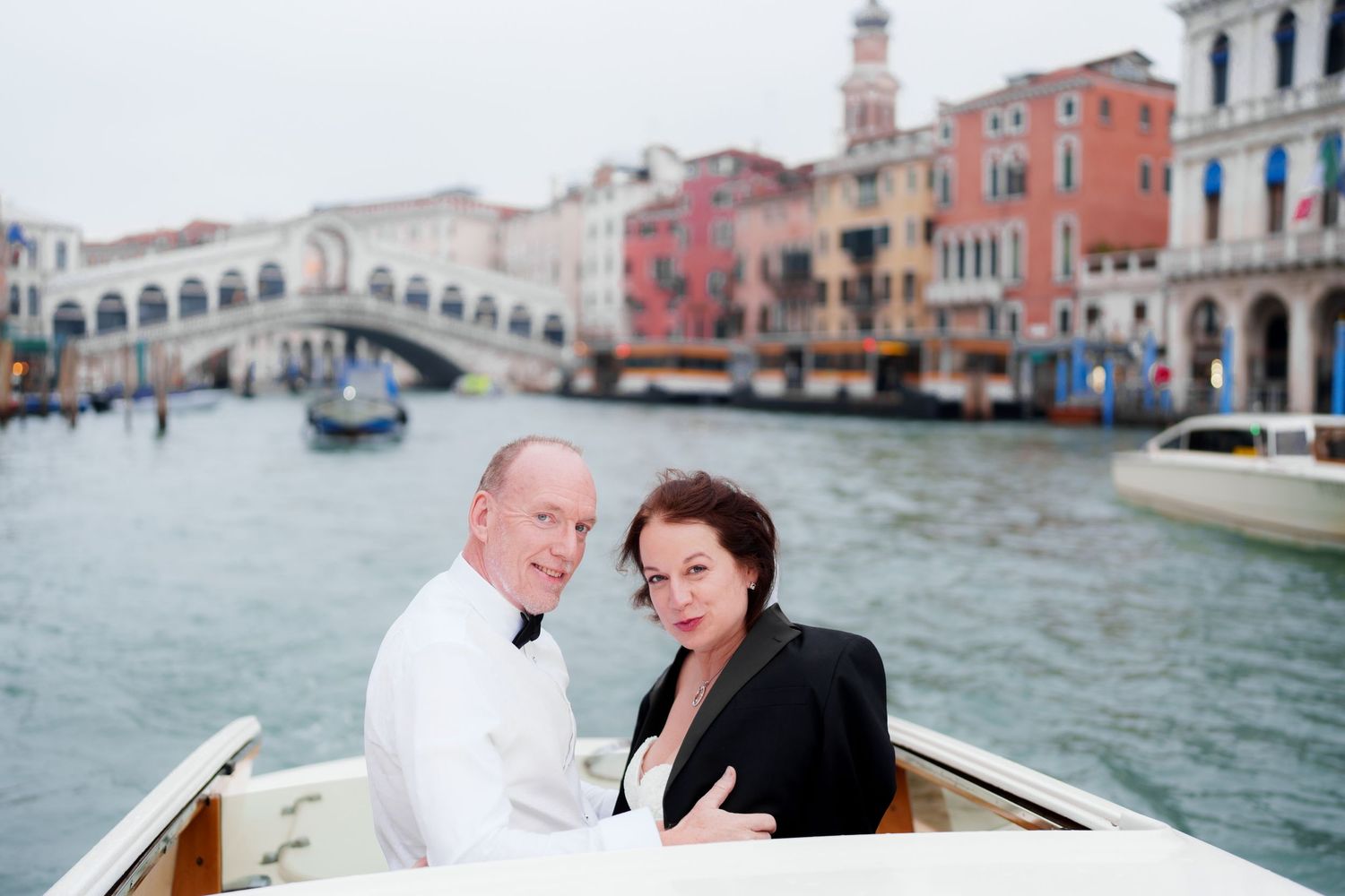 Two people enjoy a romantic boat ride along Venice's Grand Canal with the Rialto Bridge and colorful buildings in the background.
