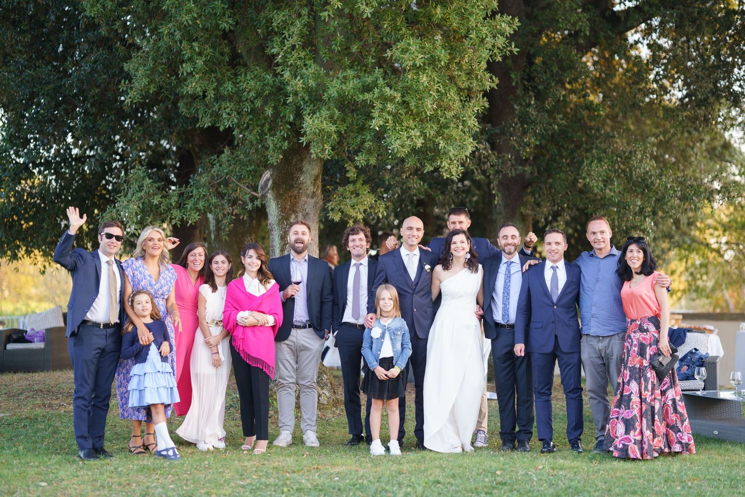 Wedding party and guests lined up for formal group photo outside with trees in background.