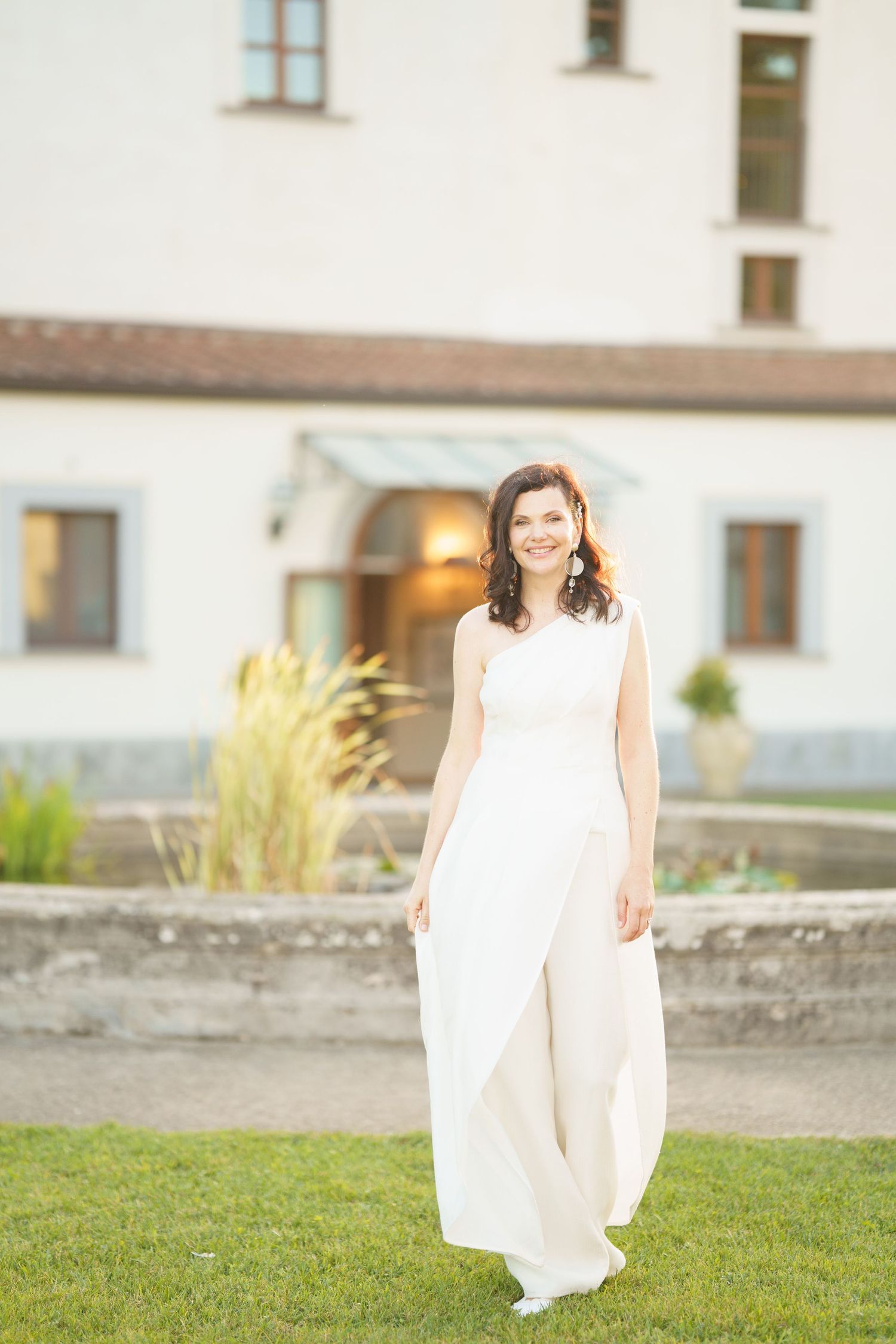 Ethereal bridal portrait on grass with white building and ornamental grasses in background.
