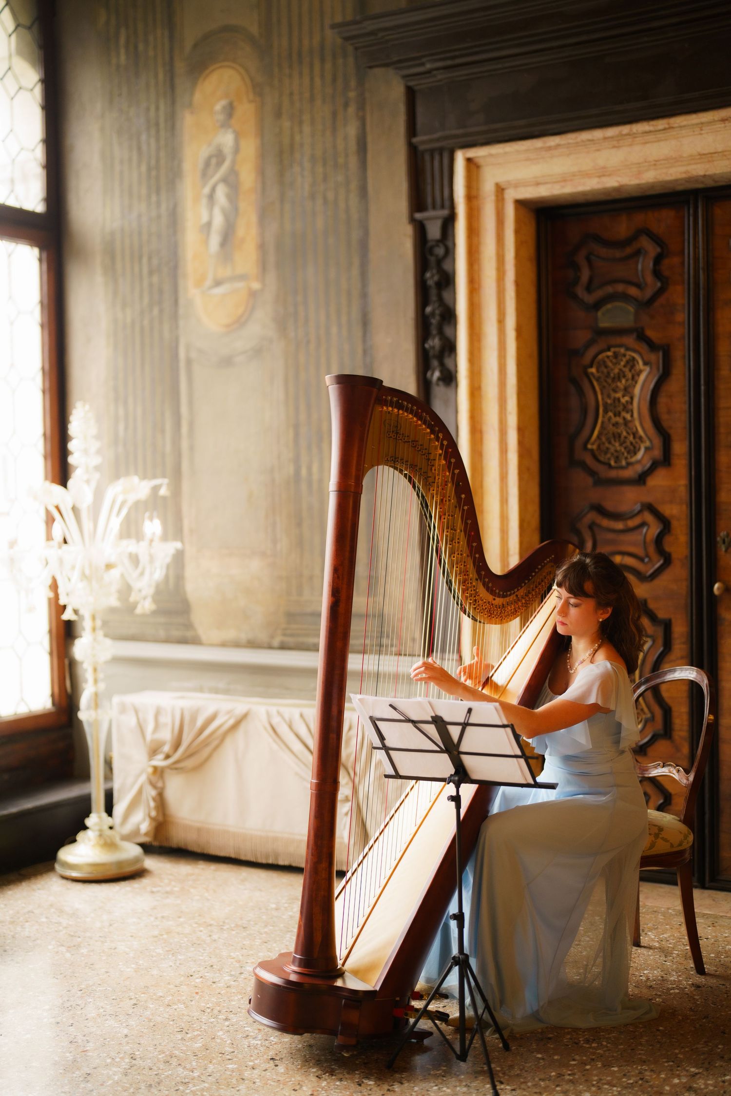 Elegant musician in white dress plays classical harp by sunlit window in ornate vintage interior.