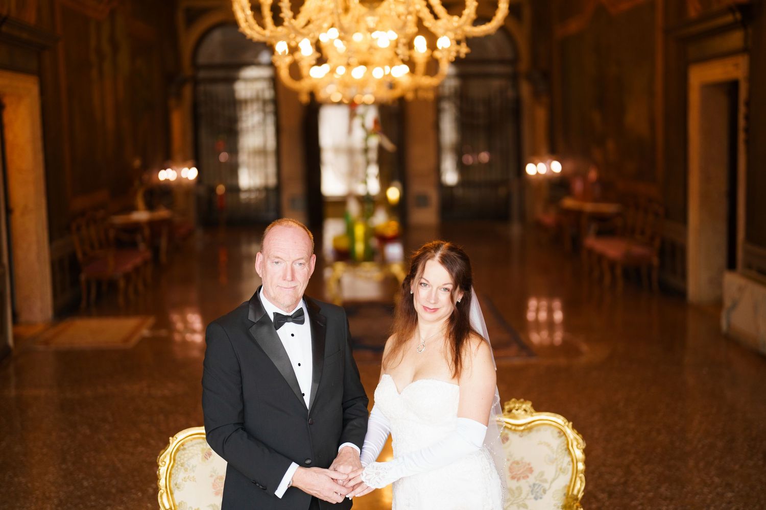 Elegant wedding couple stands beneath ornate crystal chandeliers in a grand ballroom.