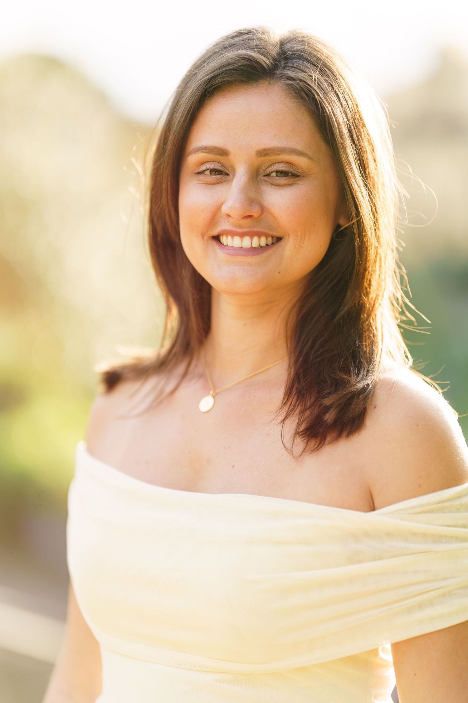 Smiling young woman with dark hair wearing an off-shoulder yellow dress and gold necklace outdoors.