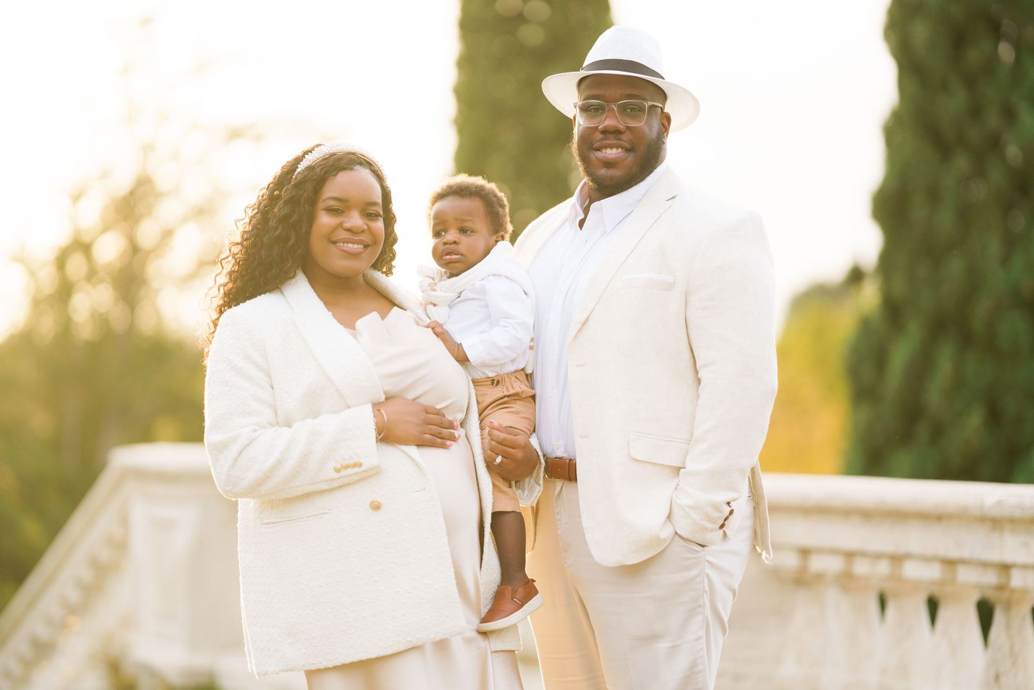 Family portrait of parents in white outfits holding baby outdoors in golden hour light.