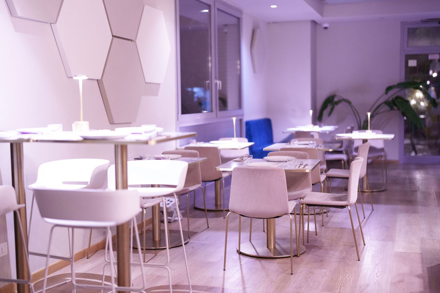 Modern white restaurant interior with hexagonal wall decor, white chairs, and gold-accented tables under purple lighting.
