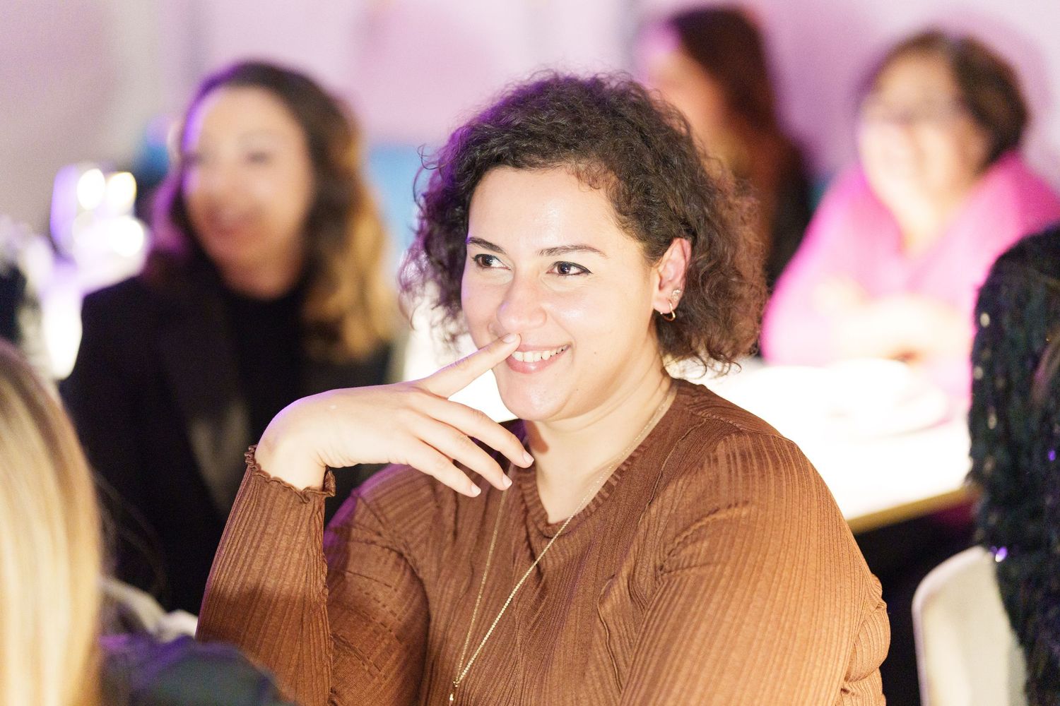 A smiling woman with curly hair in a brown top at a social event, holding a drink.