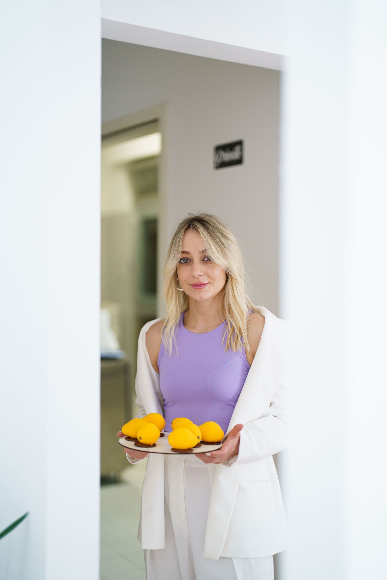 Blonde woman in white blazer and purple top holding a tray of fresh yellow lemons in a bright white interior.