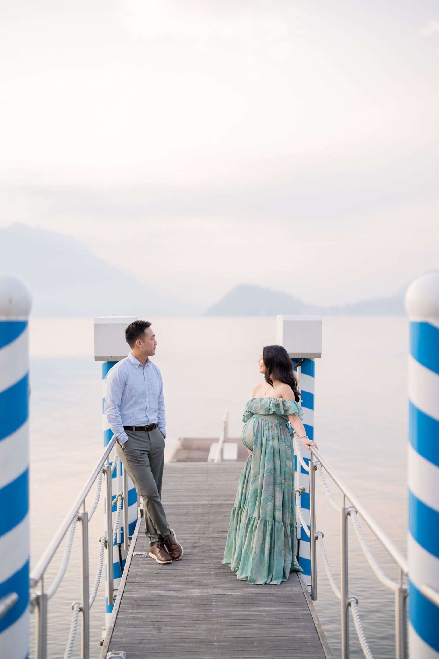 Couple standing on a blue and white pier overlooking a calm lake with mountains in the background.