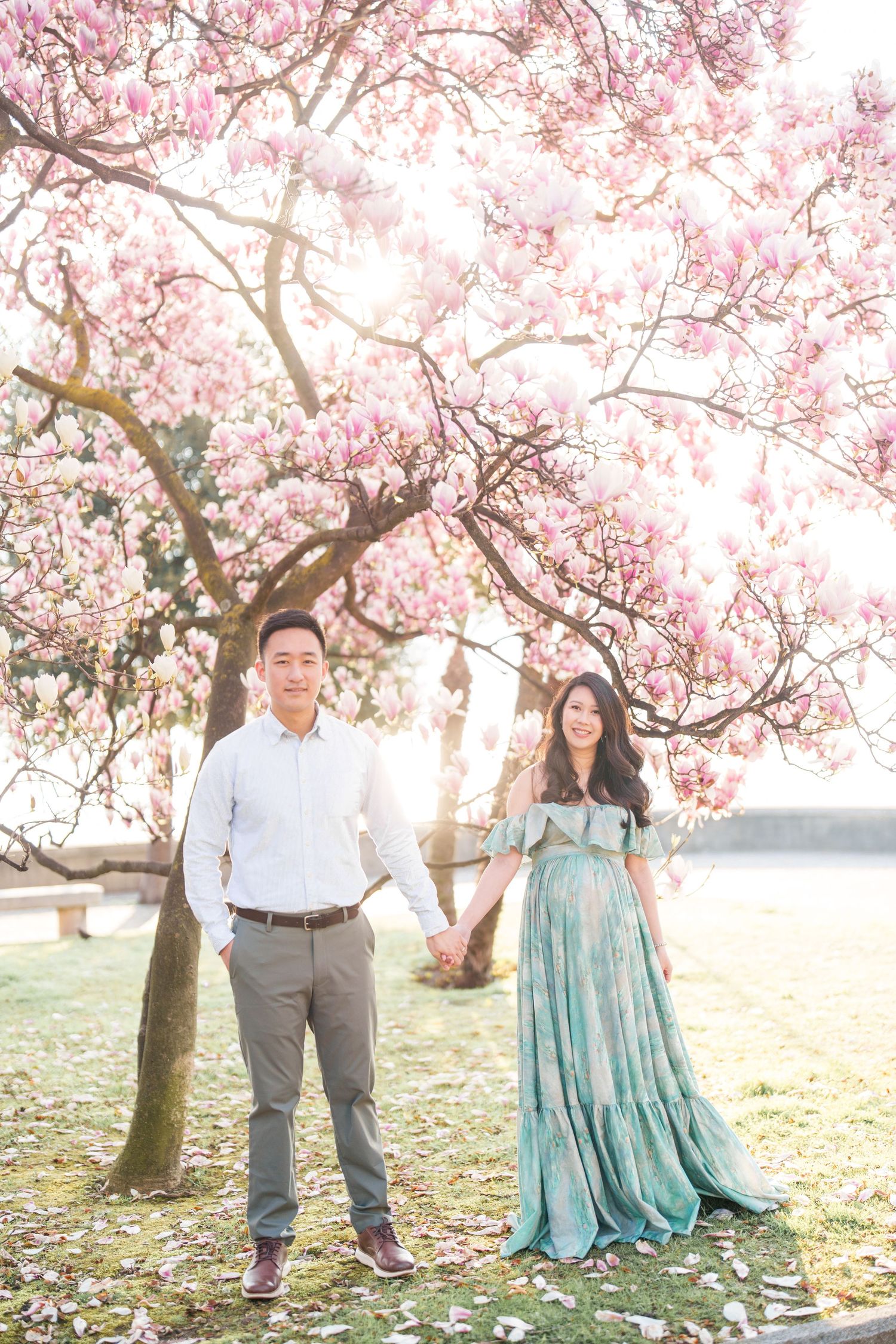 Couple holding hands under blooming pink magnolia tree, woman in teal maternity gown, man in white shirt and gray pants.