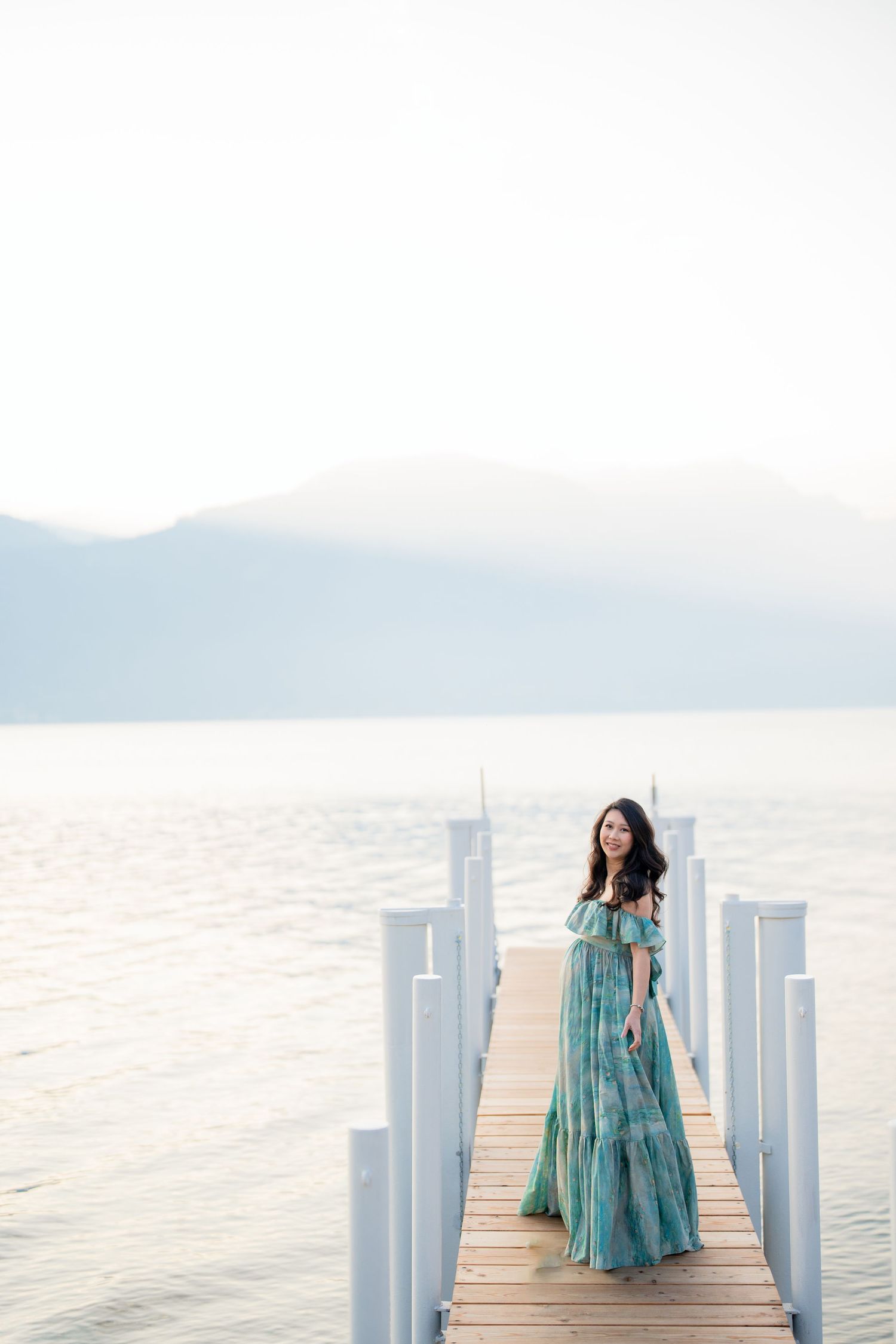 Woman in teal maxi dress standing on wooden pier overlooking calm lake with misty mountains in background.