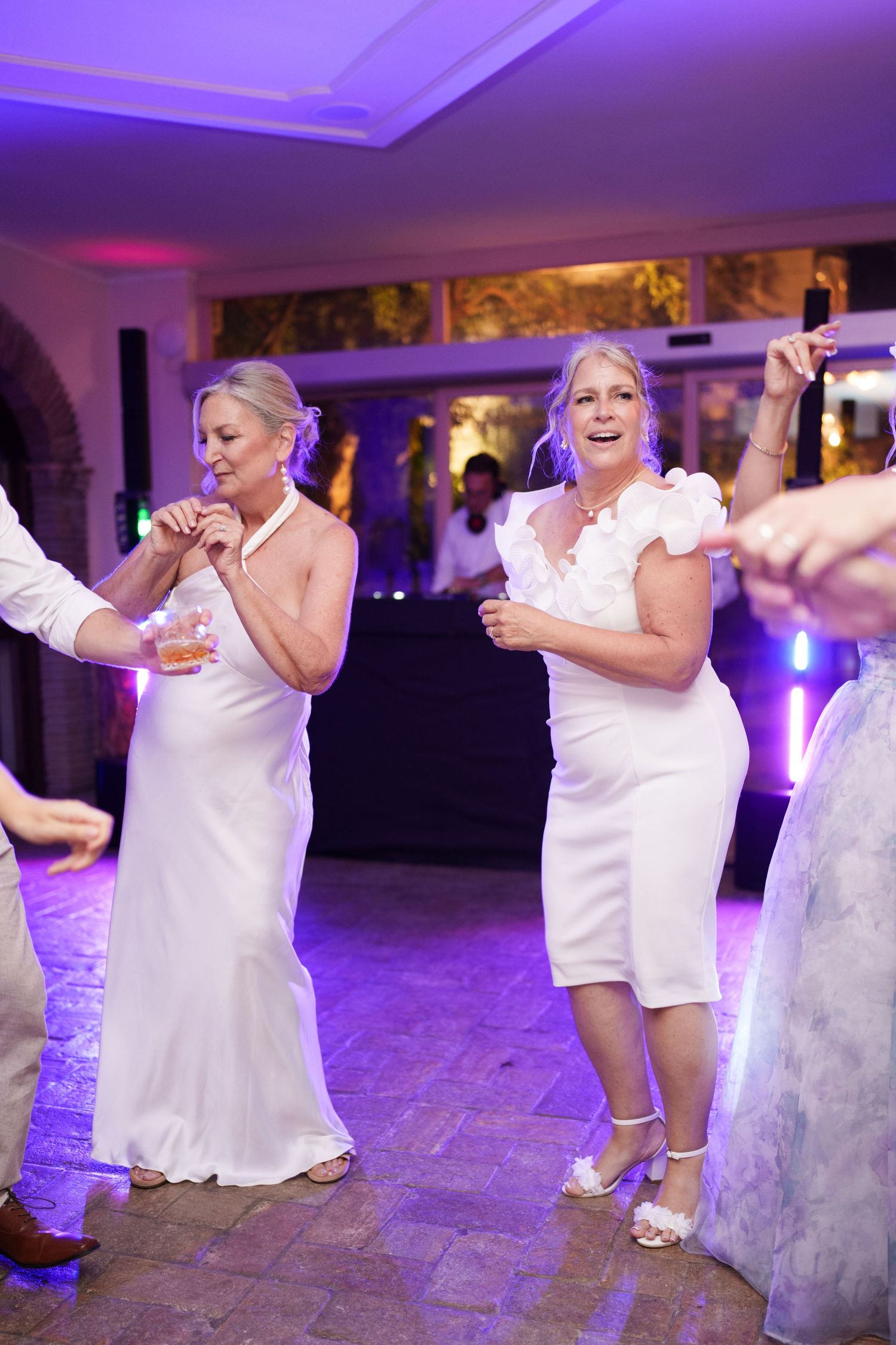 Two women in white dresses dancing joyfully at a wedding reception with purple ambient lighting.