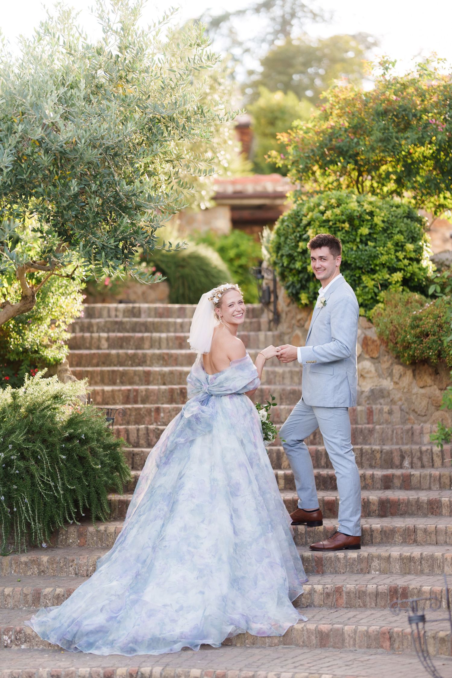 Bride in blue floral gown holds groom's hand on stone steps surrounded by lush greenery at outdoor wedding.