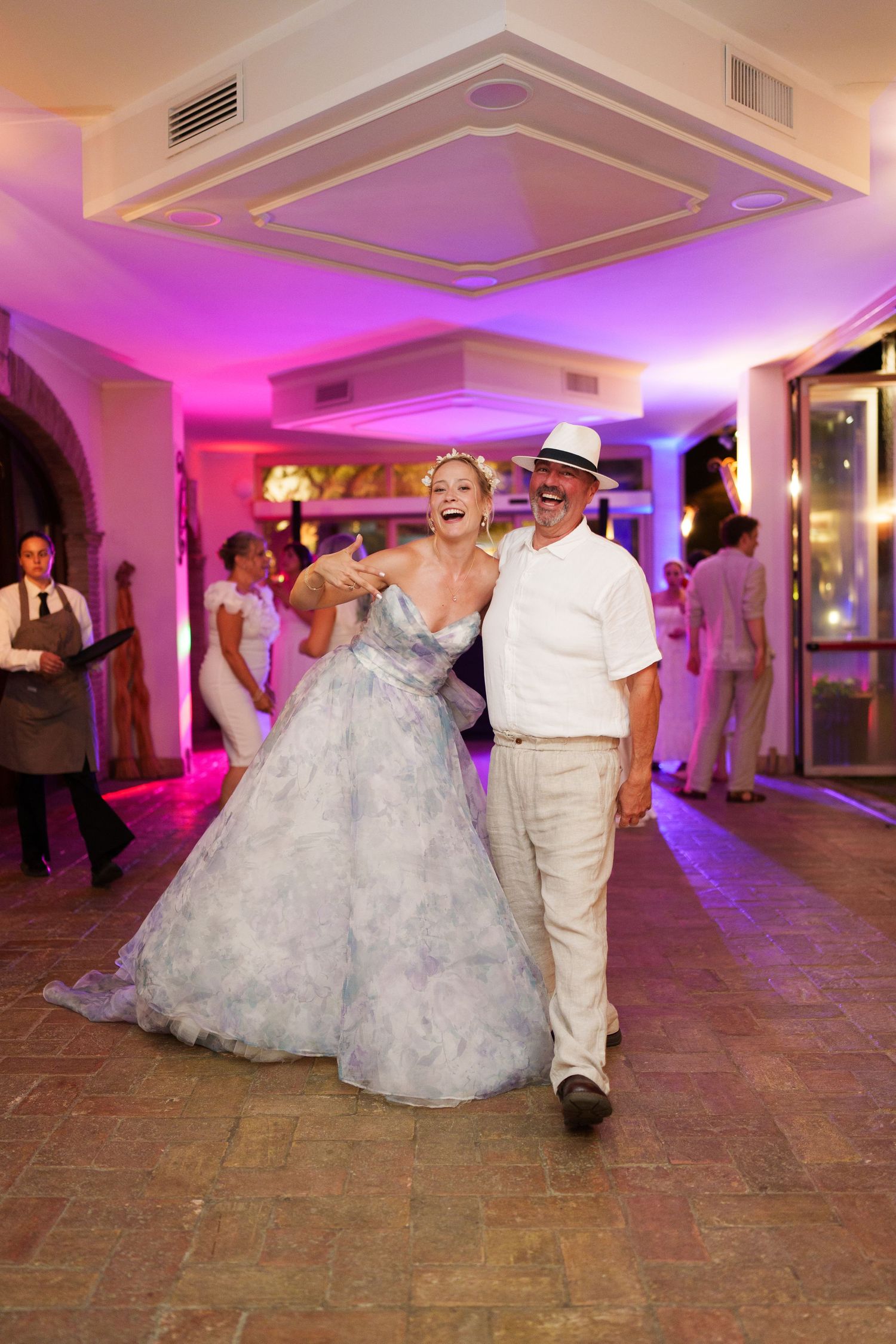 Bride in blue gown laughing with man in white suit and hat on wedding reception dance floor.