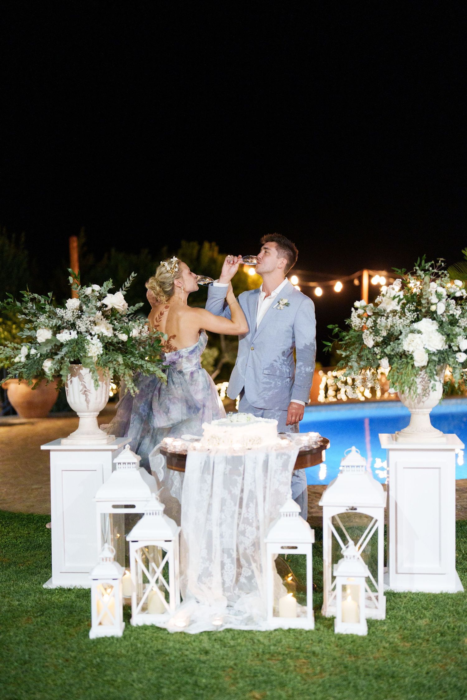 Bride and groom toasting at elegant outdoor night wedding reception with white lanterns and floral arrangements.