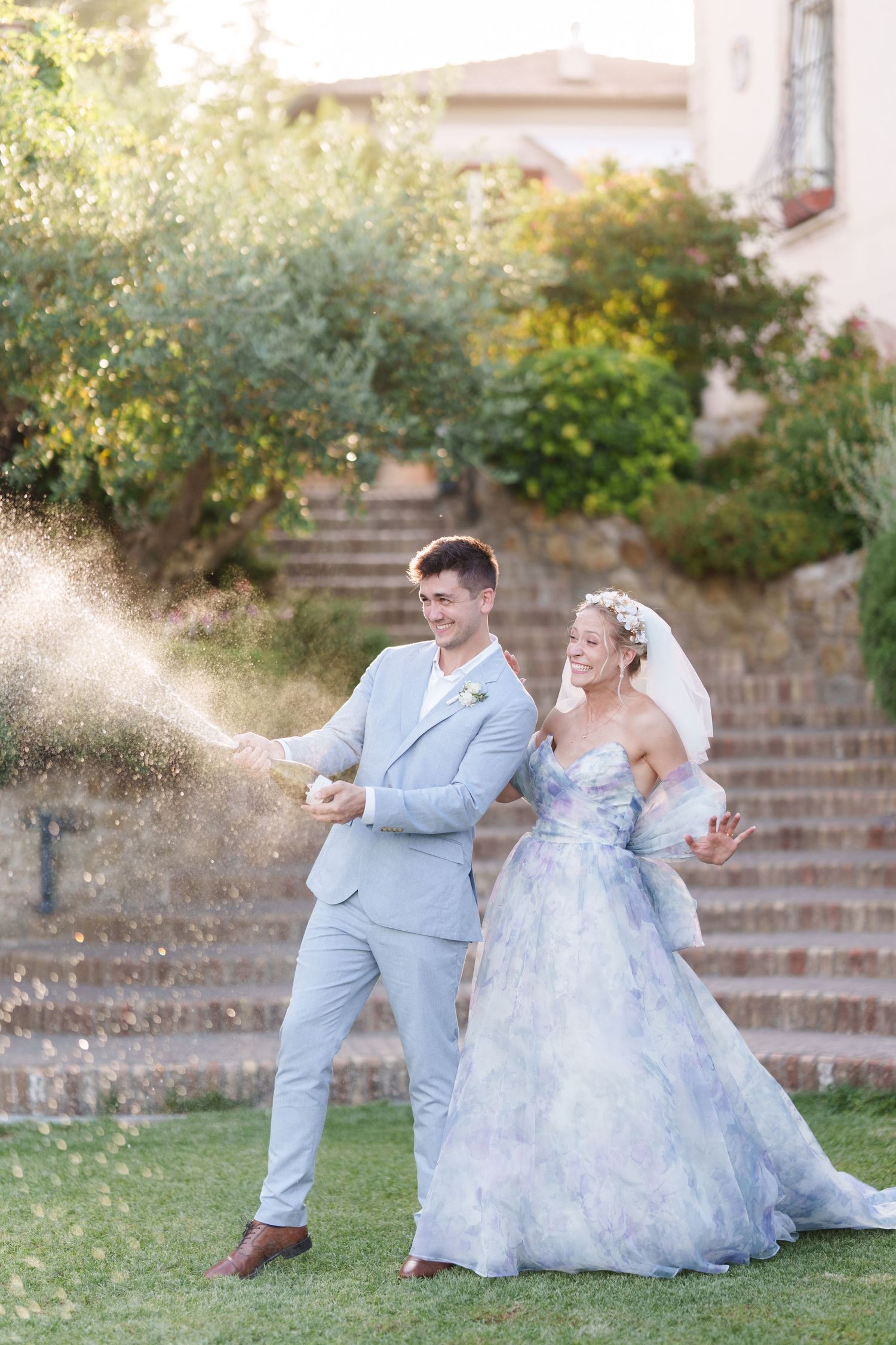 Bride and groom celebrating with champagne spray outdoors, wearing blue-gray suit and floral wedding gown.