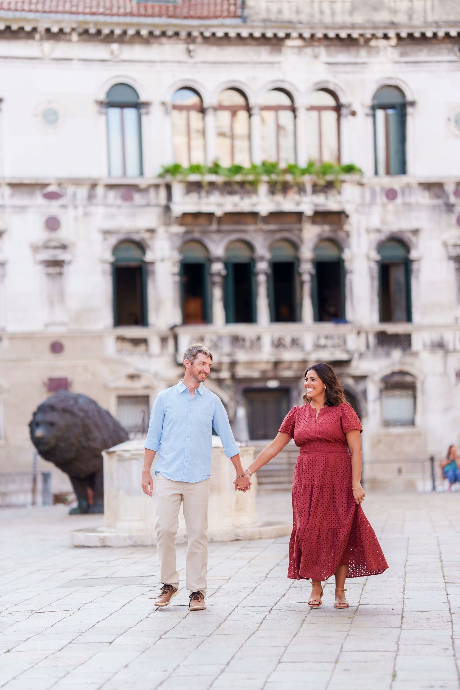 Couple holding hands and walking in a Venetian piazza with historic architecture and a lion sculpture in the background.