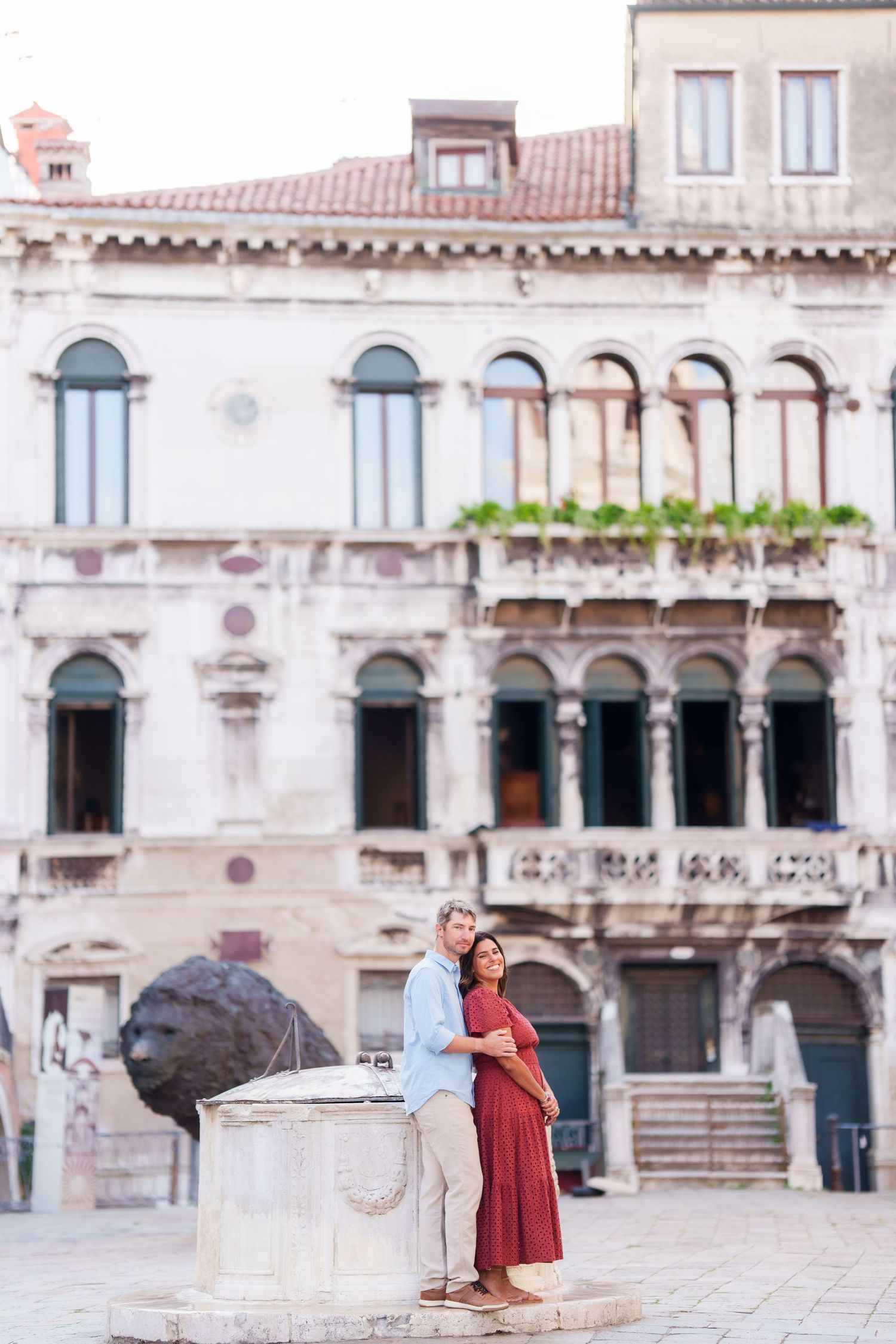 Couple embracing by a stone well in a Venetian piazza, with ornate historic architecture in the background.