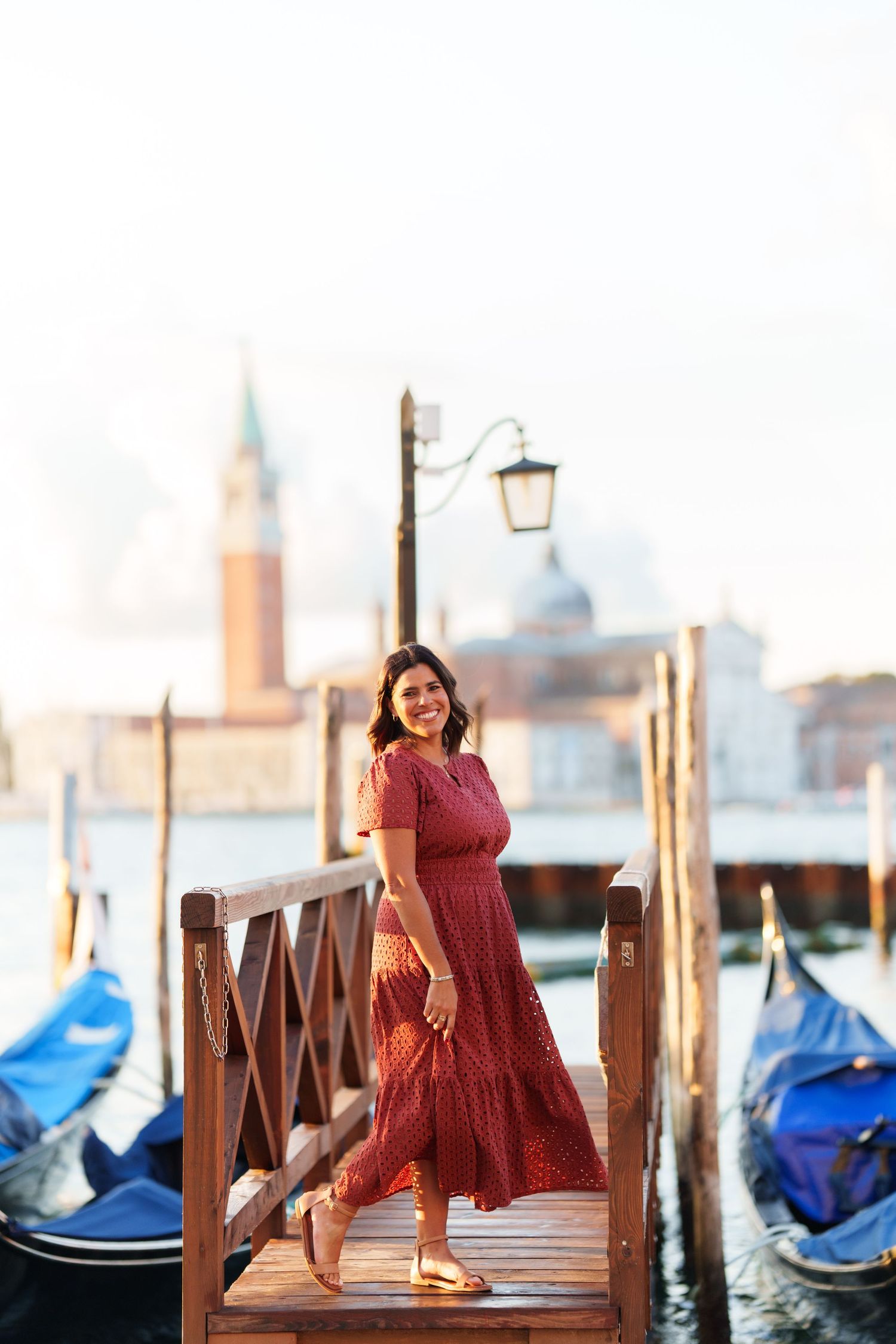 Woman in red dress smiling on Venice gondola dock with San Giorgio Maggiore church in background.