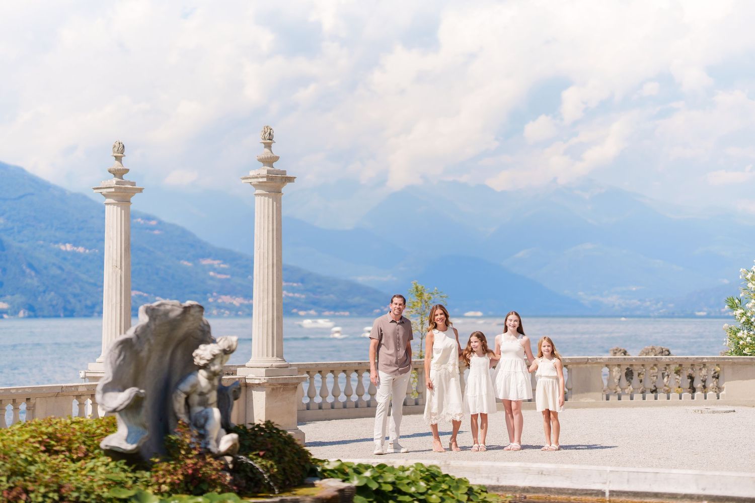 Family in white outfits posing on elegant terrace with marble columns, Lake Como and mountain backdrop.