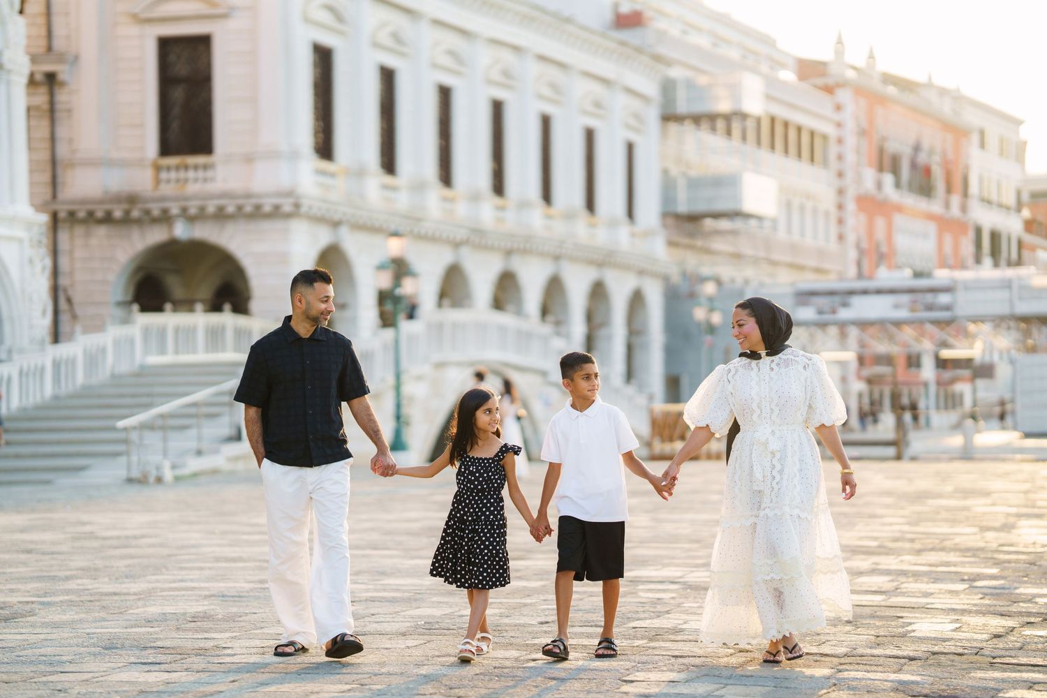 Family of four holding hands and walking in a sunlit European plaza with ornate architecture.