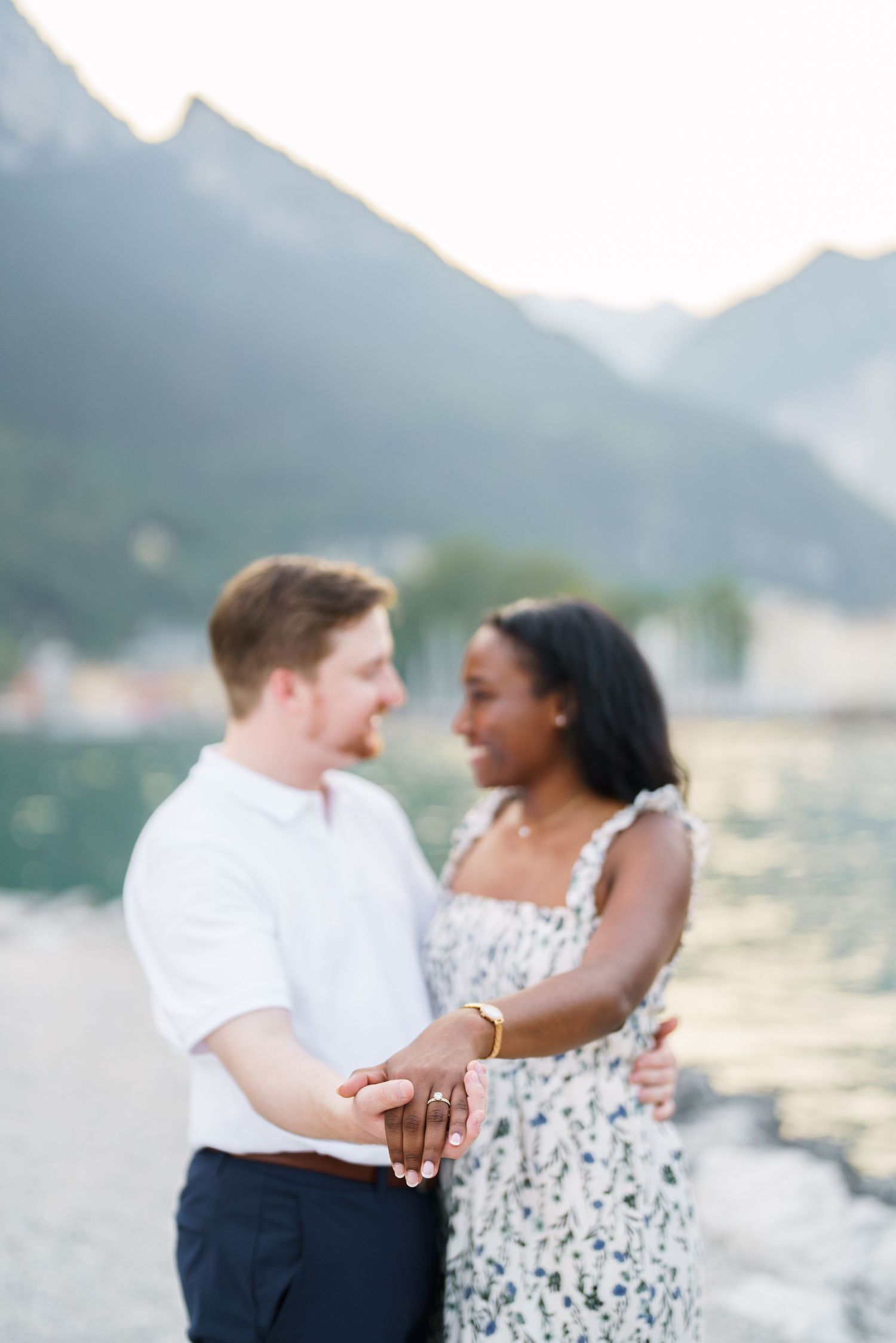 Engaged interracial couple holding hands and gazing at each other by a sunlit mountain lake.