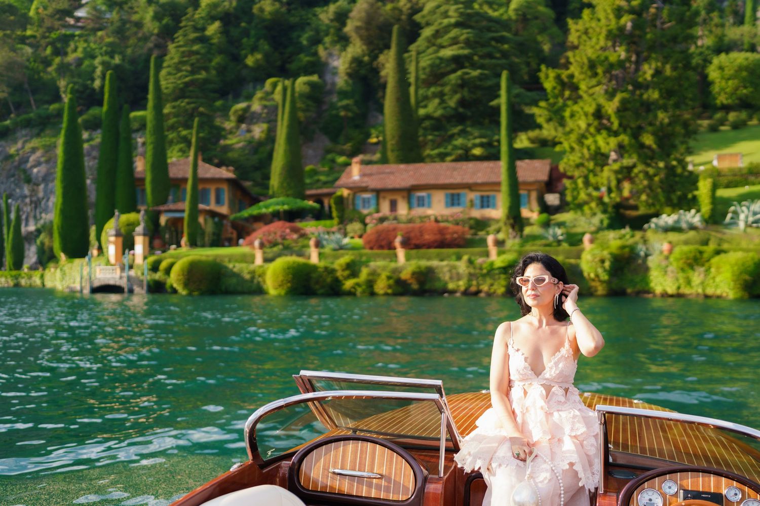 Elegant woman in white lace dress on a wooden boat at Lake Como, with lush greenery and villa in background.