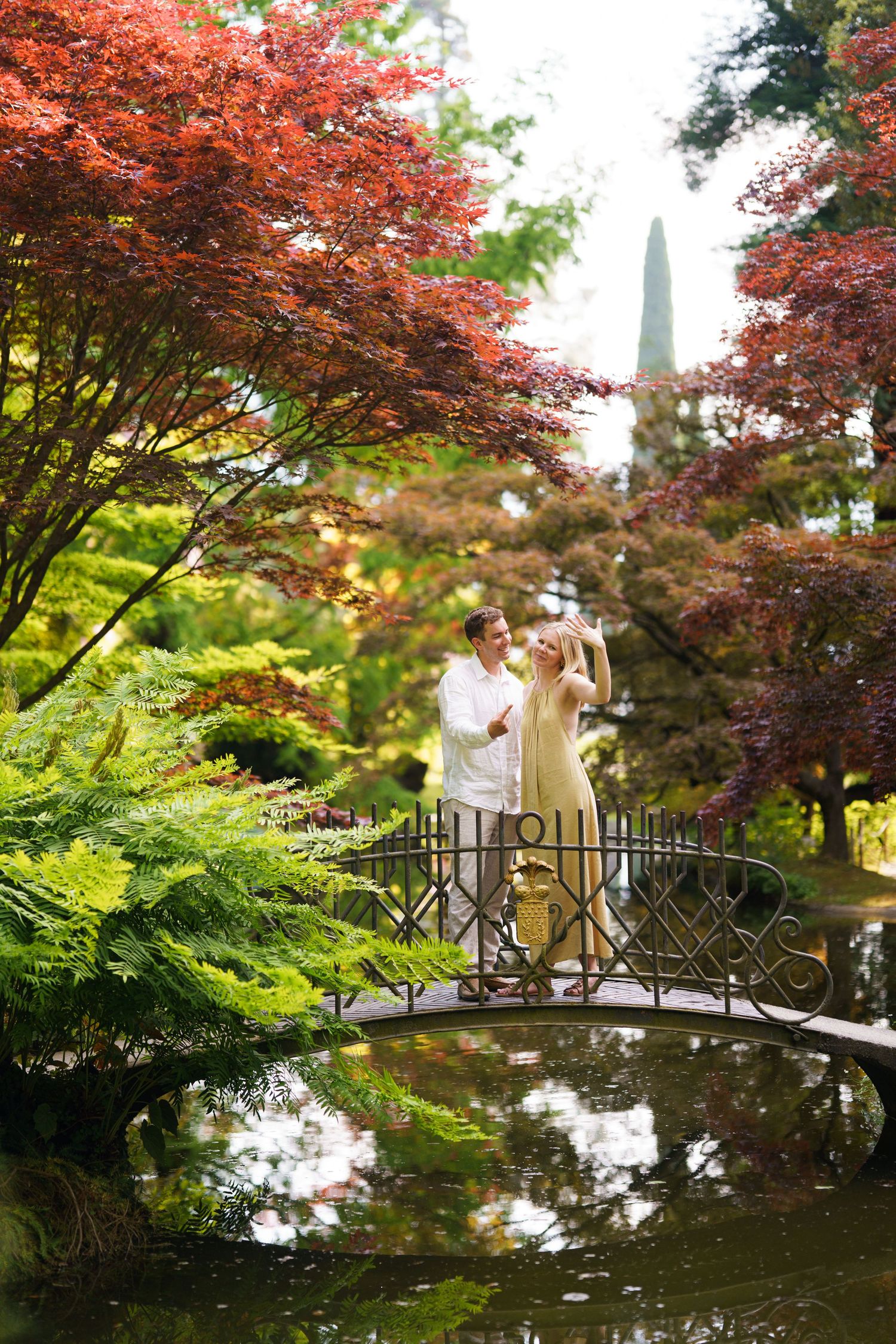 Couple dancing on ornate iron bridge over pond in lush Japanese garden with vibrant red maple trees.