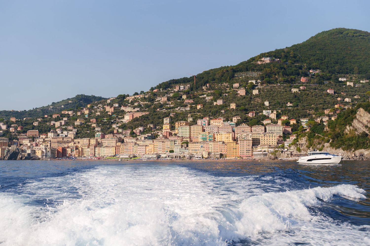 Colorful Italian coastal village with lush green hills, viewed from a speedboat leaving white wake on blue sea.