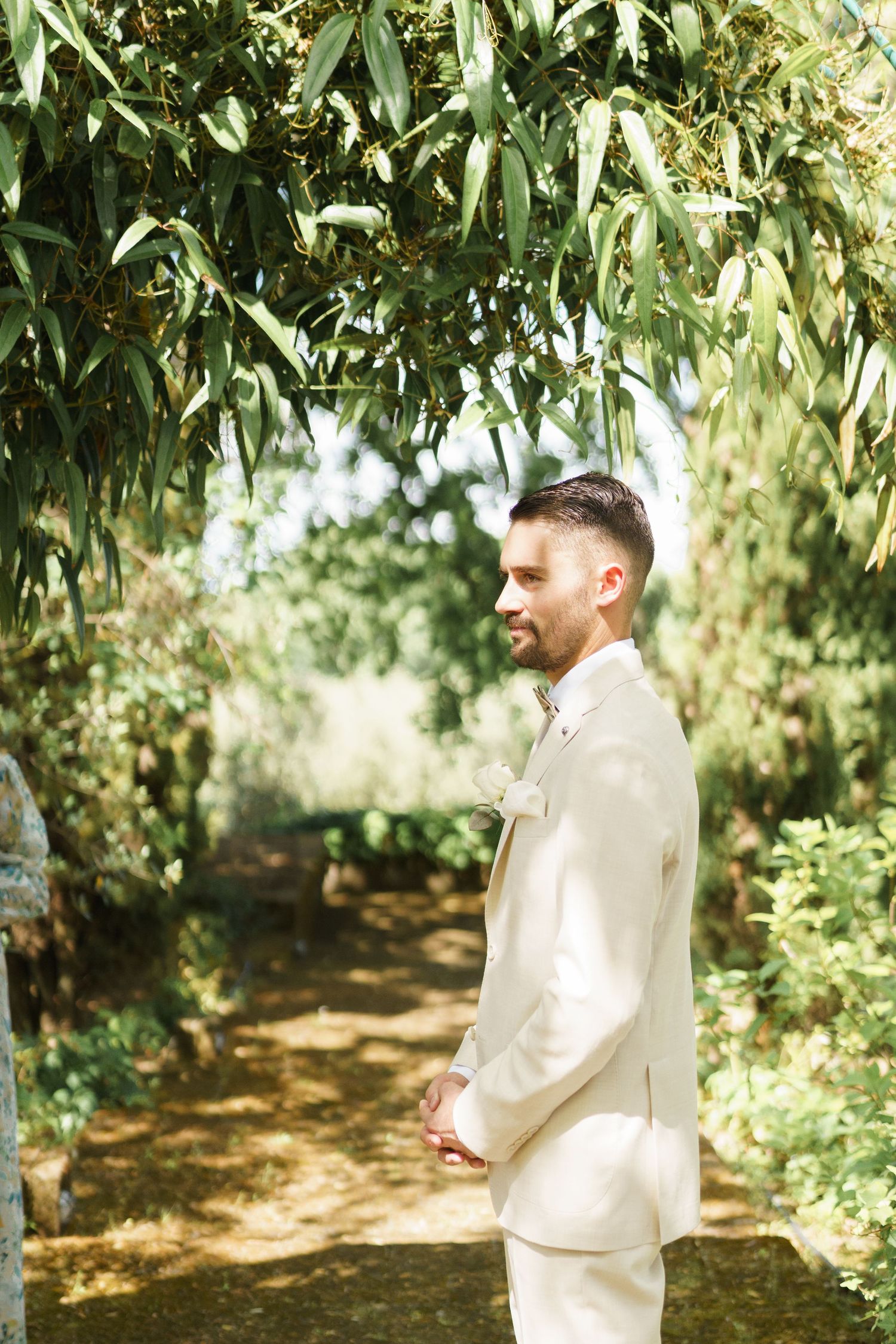 Groom in cream suit standing in lush garden path, smiling profile view with green trees overhead on sunny day.