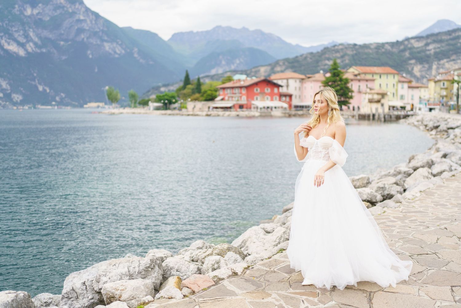 Bride in white gown poses on rocky lakeside with mountain village backdrop on a cloudy day.