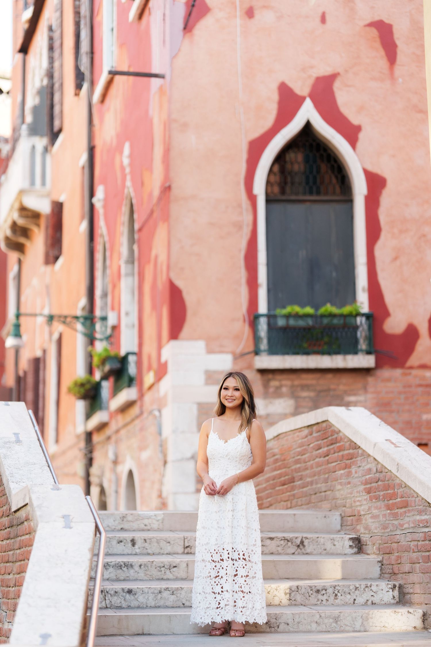 Woman in white lace dress standing on marble steps before colorful Venetian buildings in Venice, Italy.