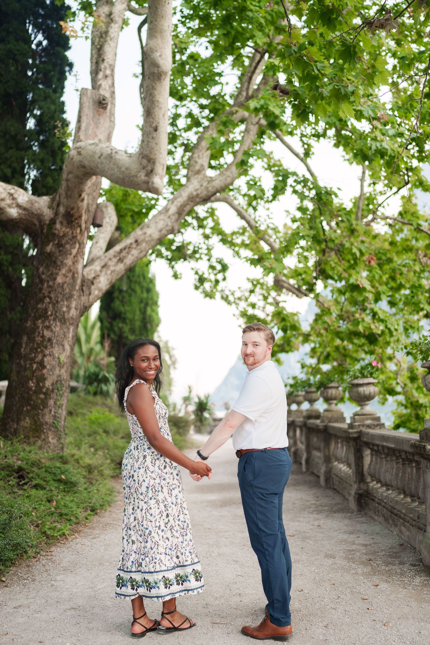 Smiling couple holding hands on a tree-lined garden path with a decorative stone balustrade in Italy.
