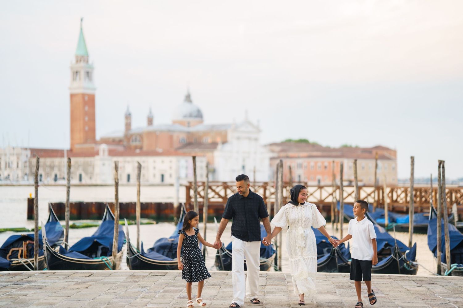 Family of four holding hands by Venice gondolas with San Giorgio Maggiore church in background.