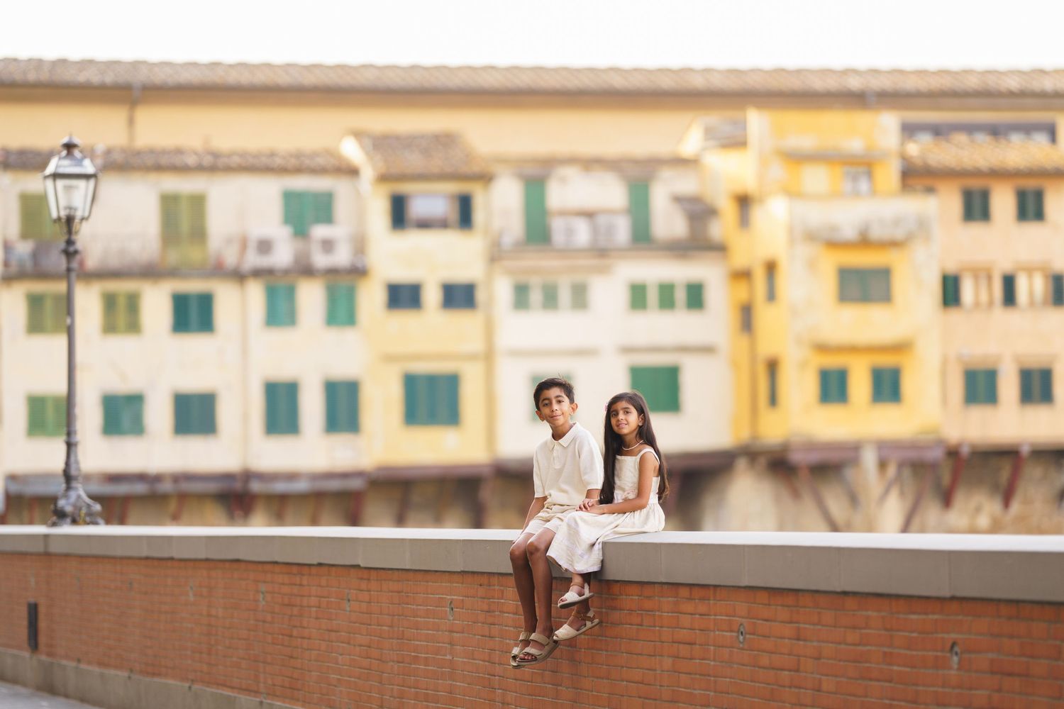 Two children in white outfits sit on a wall with colorful Ponte Vecchio buildings in Florence, Italy behind them.