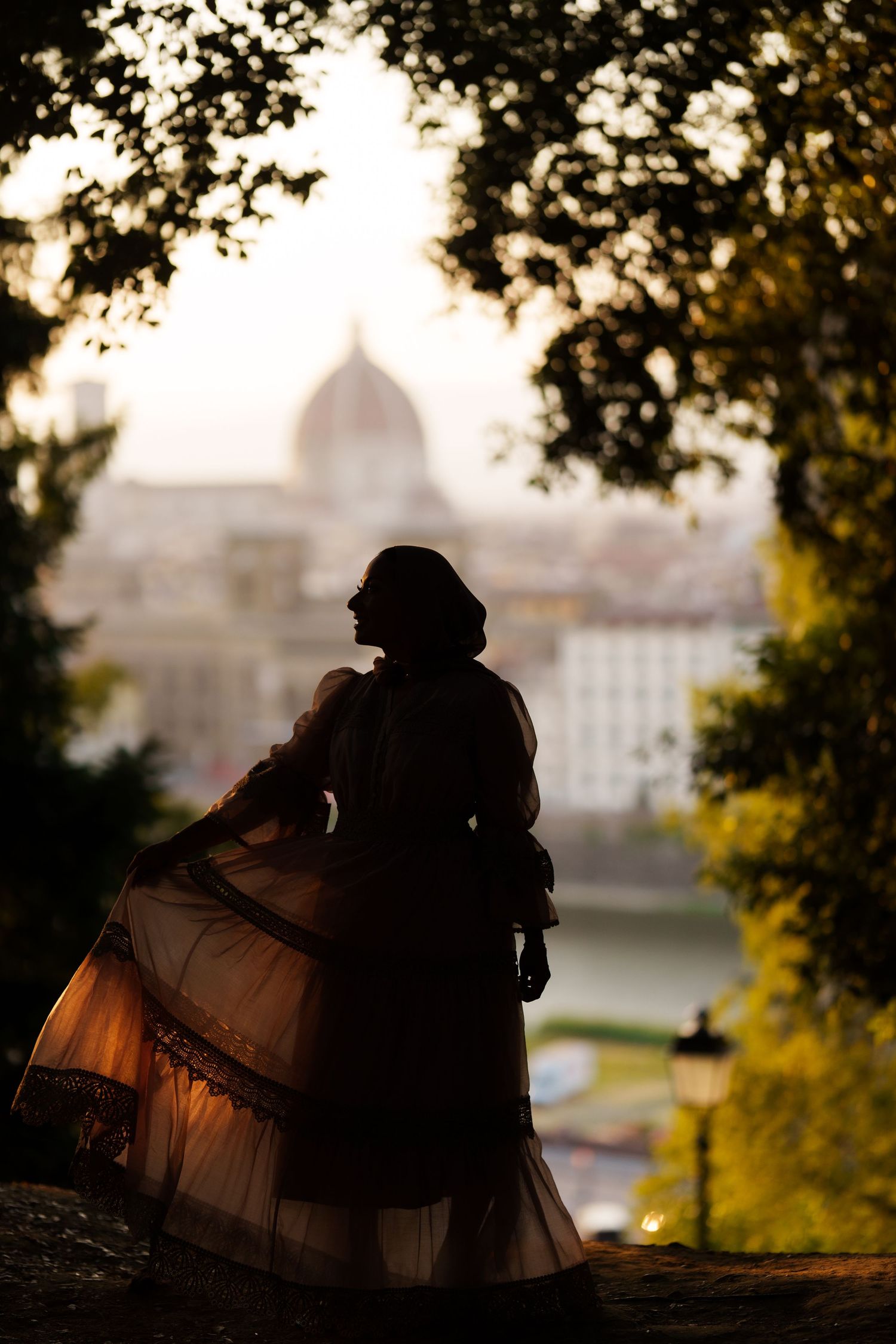 Silhouette of a woman in a flowing gown framed by trees with Florence cathedral dome at sunset.
