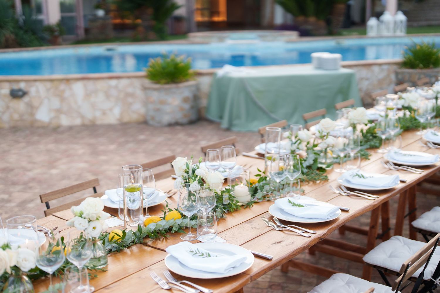 Elegant outdoor wedding reception table set by a pool with white floral centerpieces, greenery runner, and fine dining place settings.